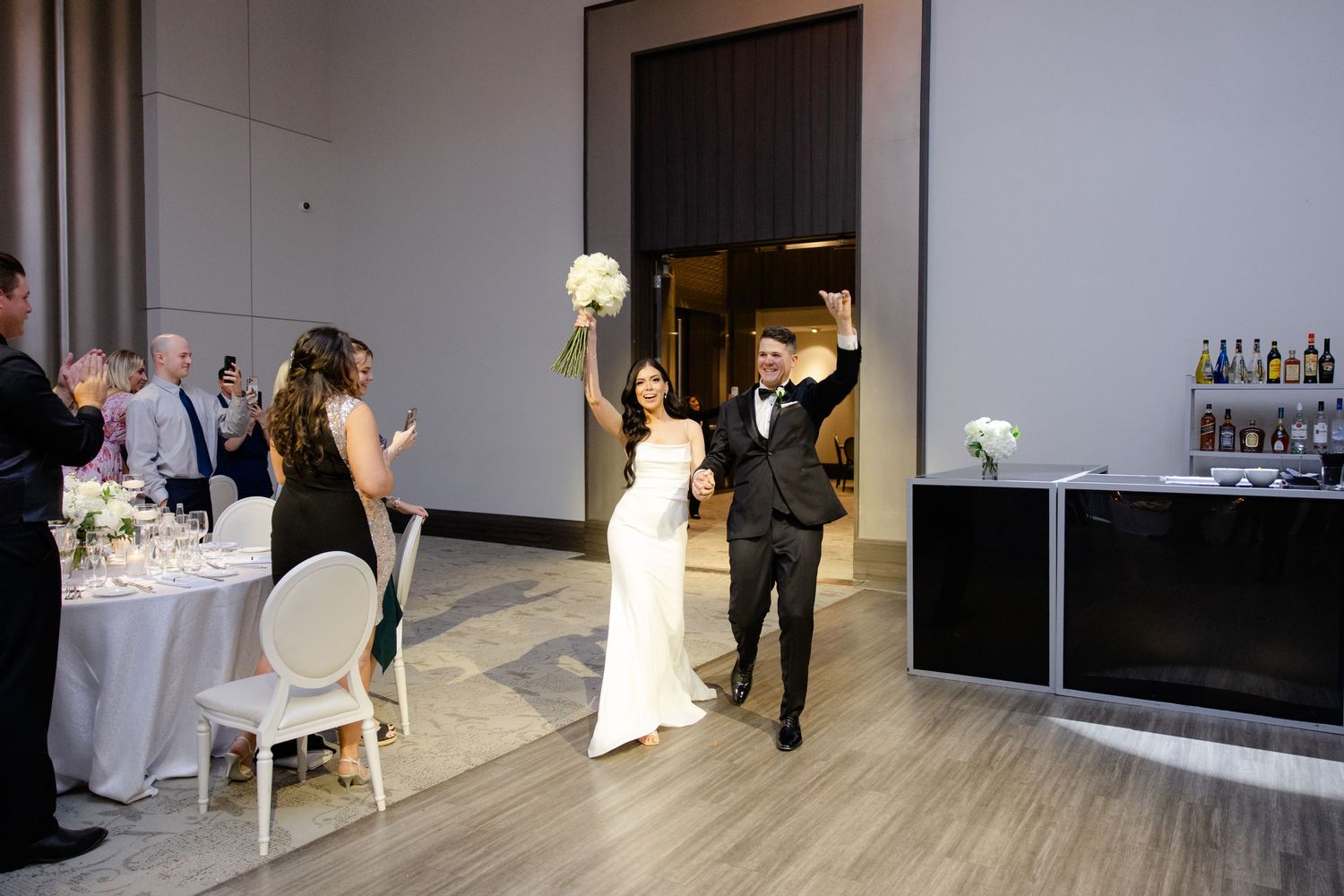Wedding couple enters reception venue as guests seated at tables applaud their arrival.