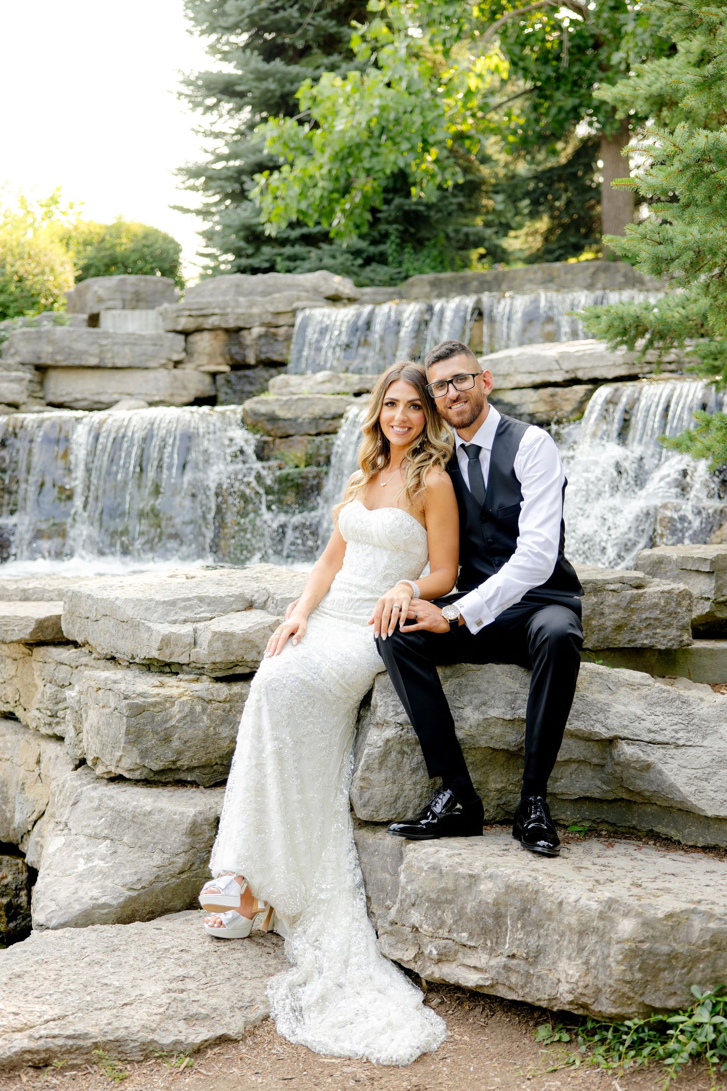 Wedding couple seated on stone waterfall steps in garden setting.