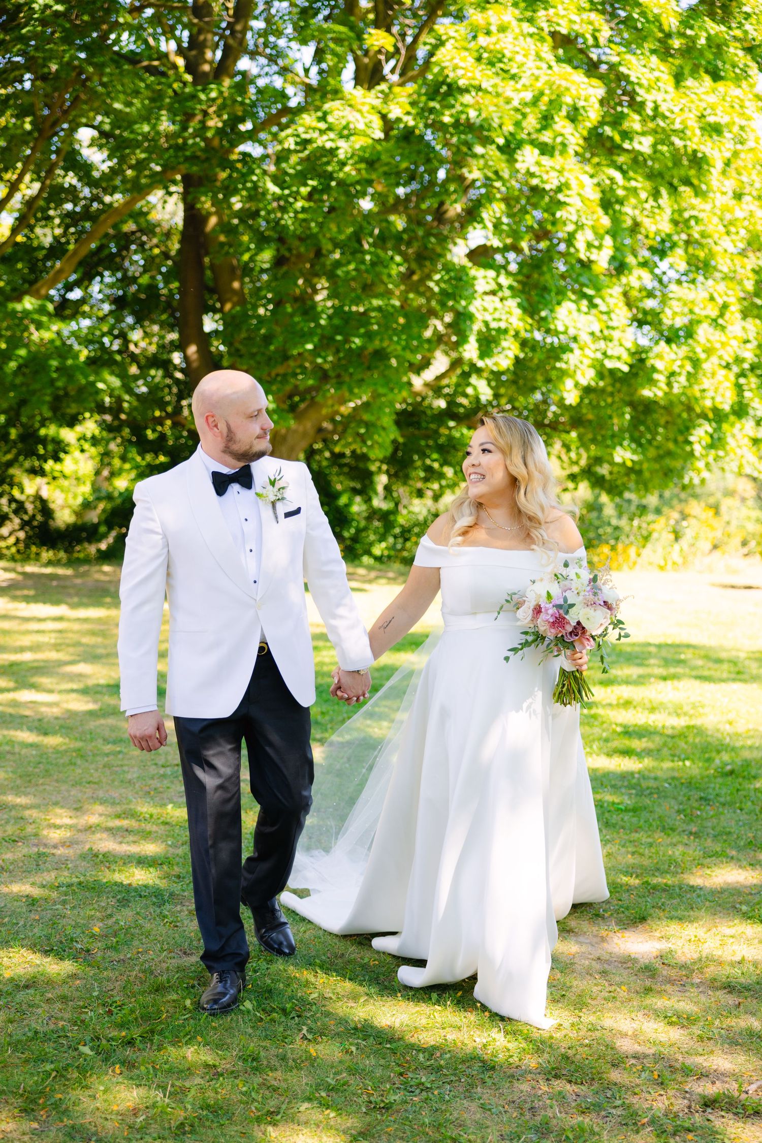 A couple walks hand in hand through a sunlit garden on their wedding day, with the bride holding a pink bouquet.