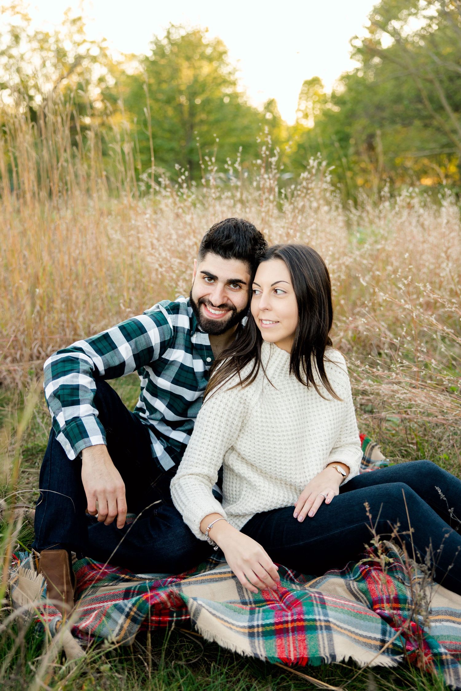 Couple sits on plaid blanket in autumn field during sunset photoshoot.
