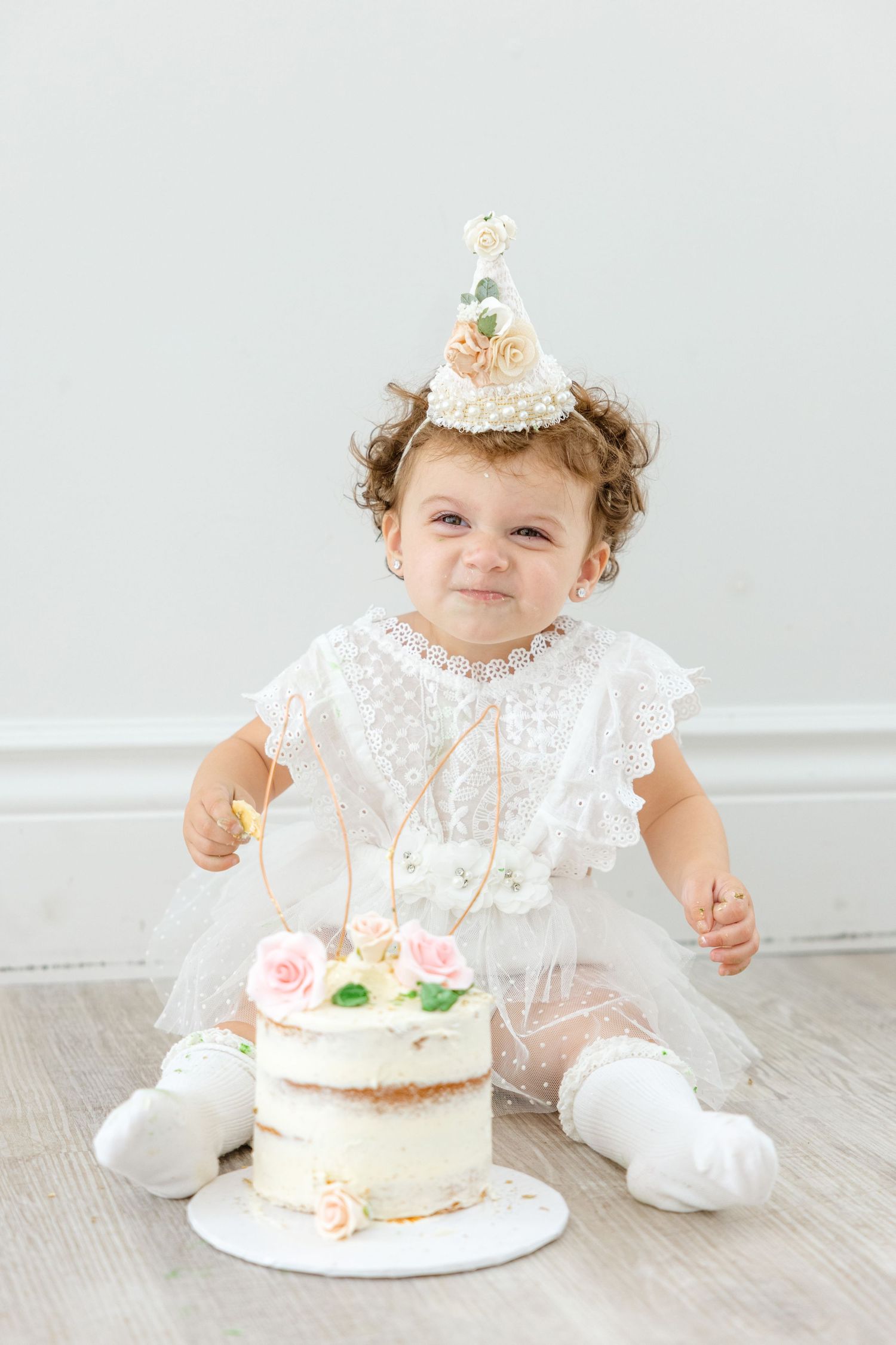 A toddler in a white dress and party hat sits with a decorated birthday cake on the floor.
