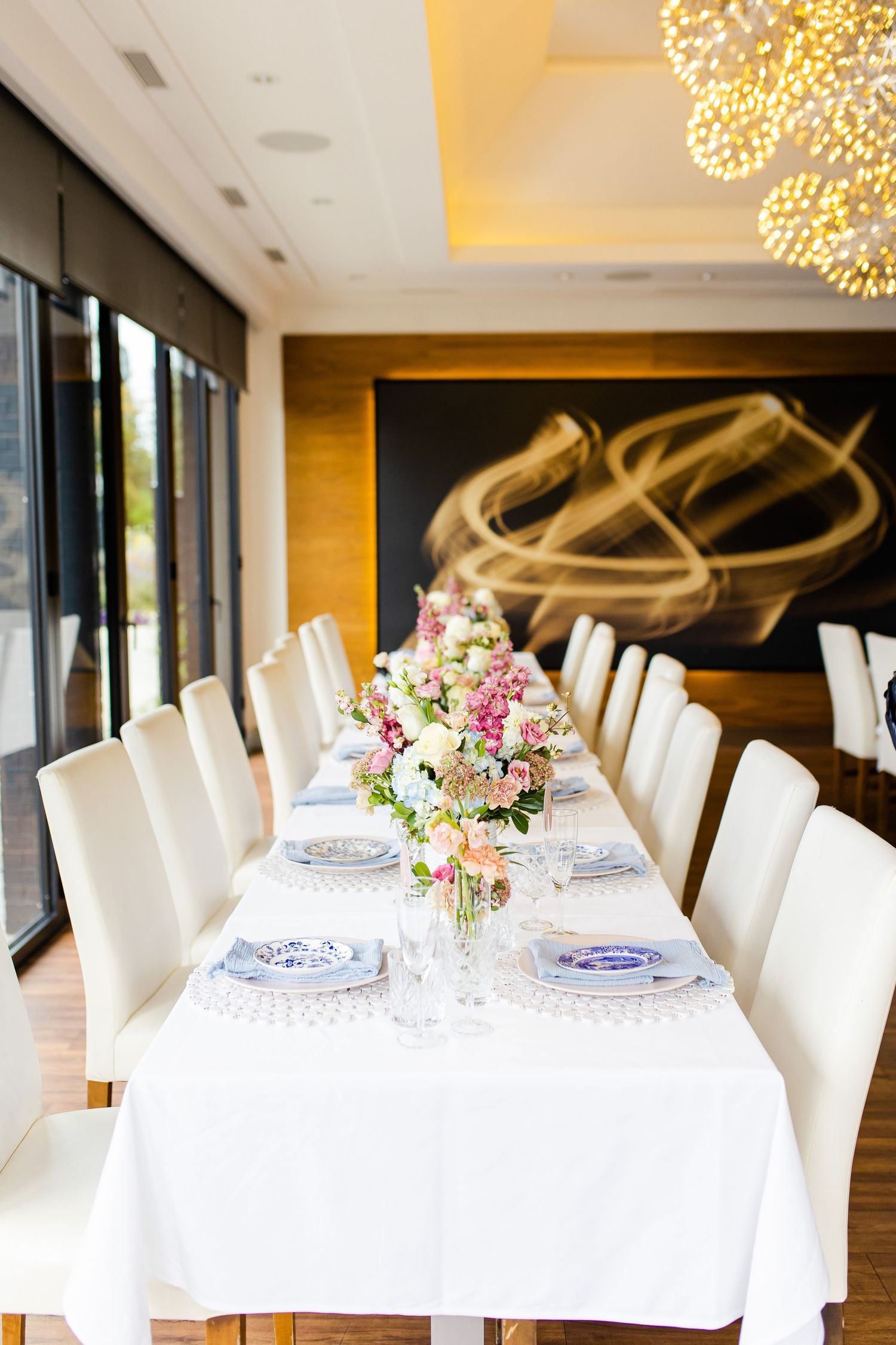 Long white dining table with cream chairs and fresh floral arrangements beneath pendant lights.