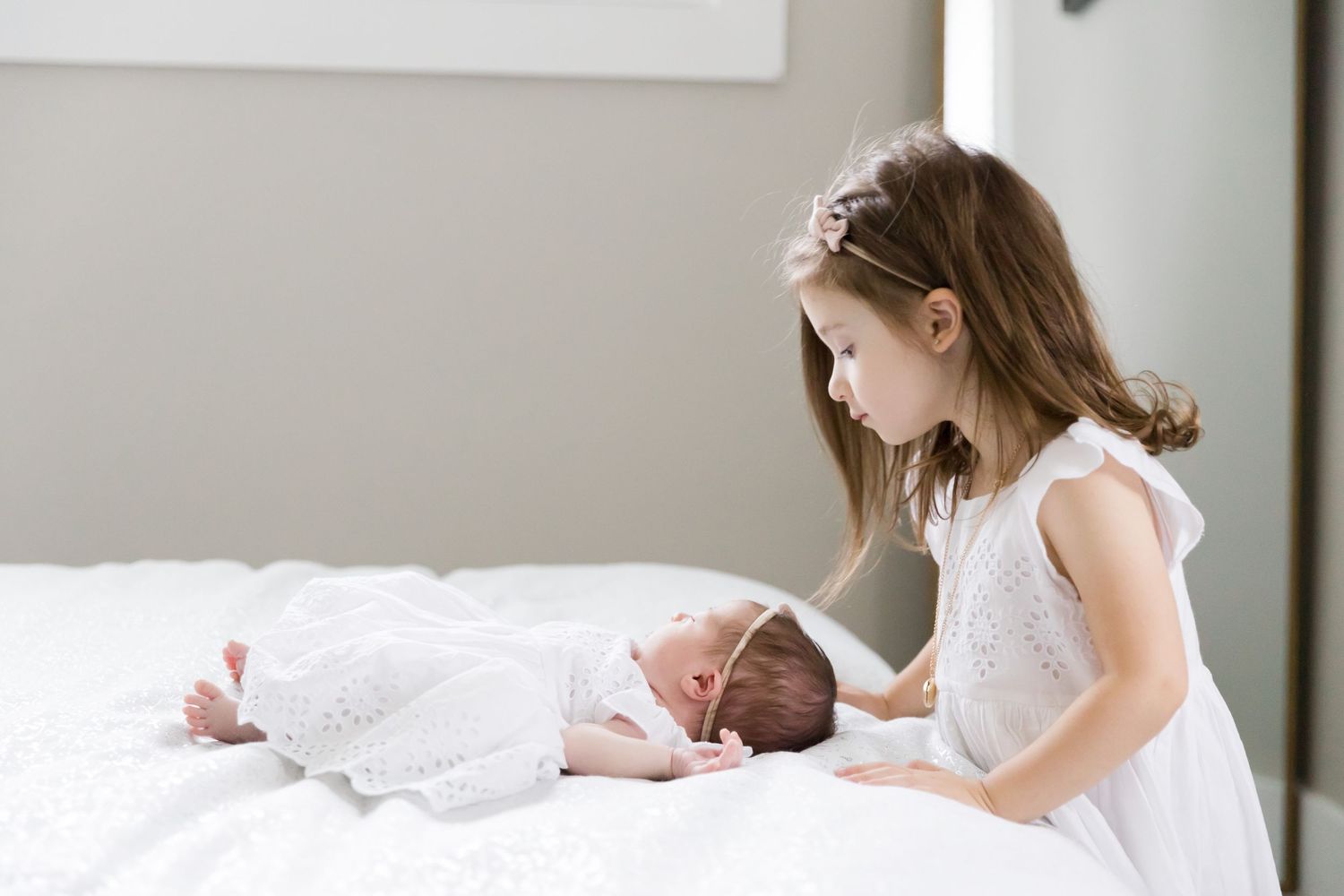 Child in white dress looking at sleeping baby on bed.