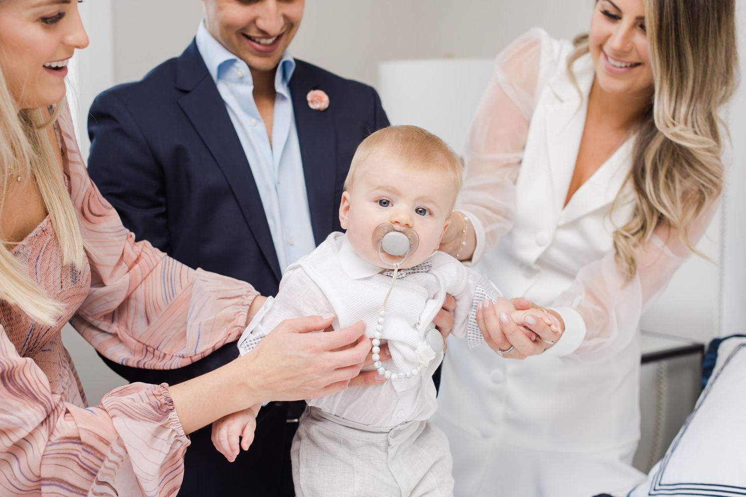 Baby with pacifier being supported by adults in formal attire.