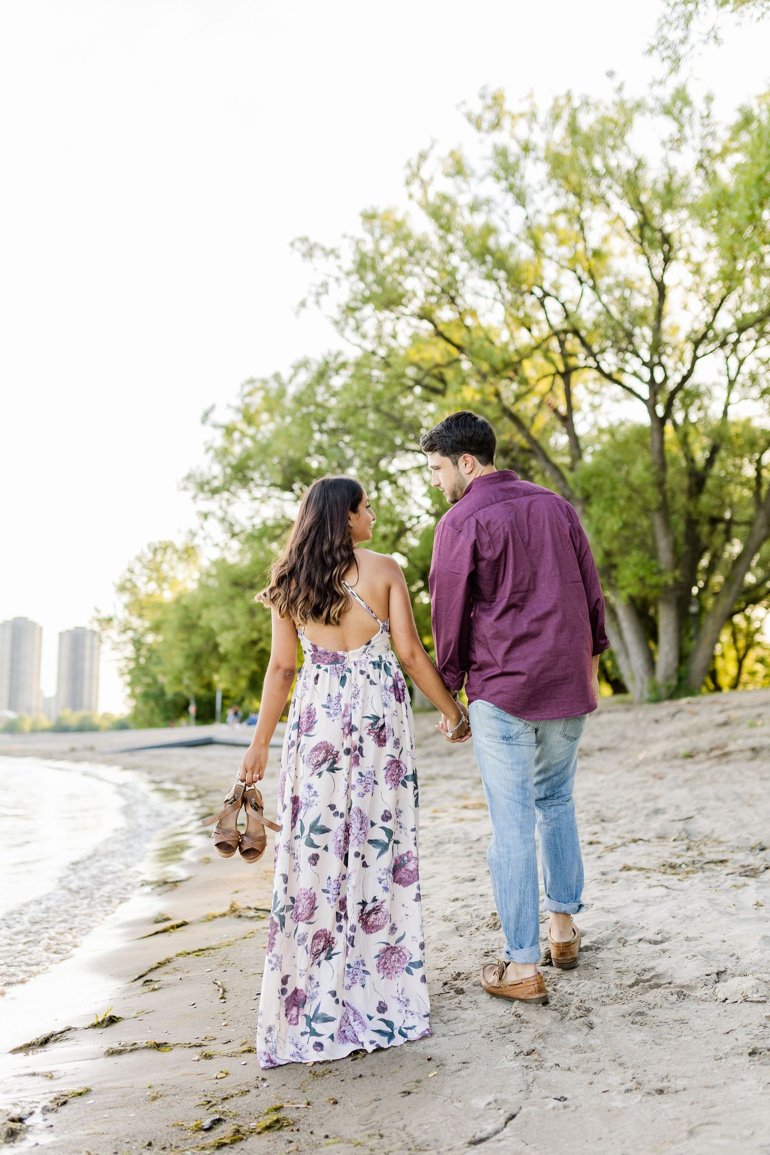 Couple walking hand-in-hand along shoreline with urban skyline visible.
