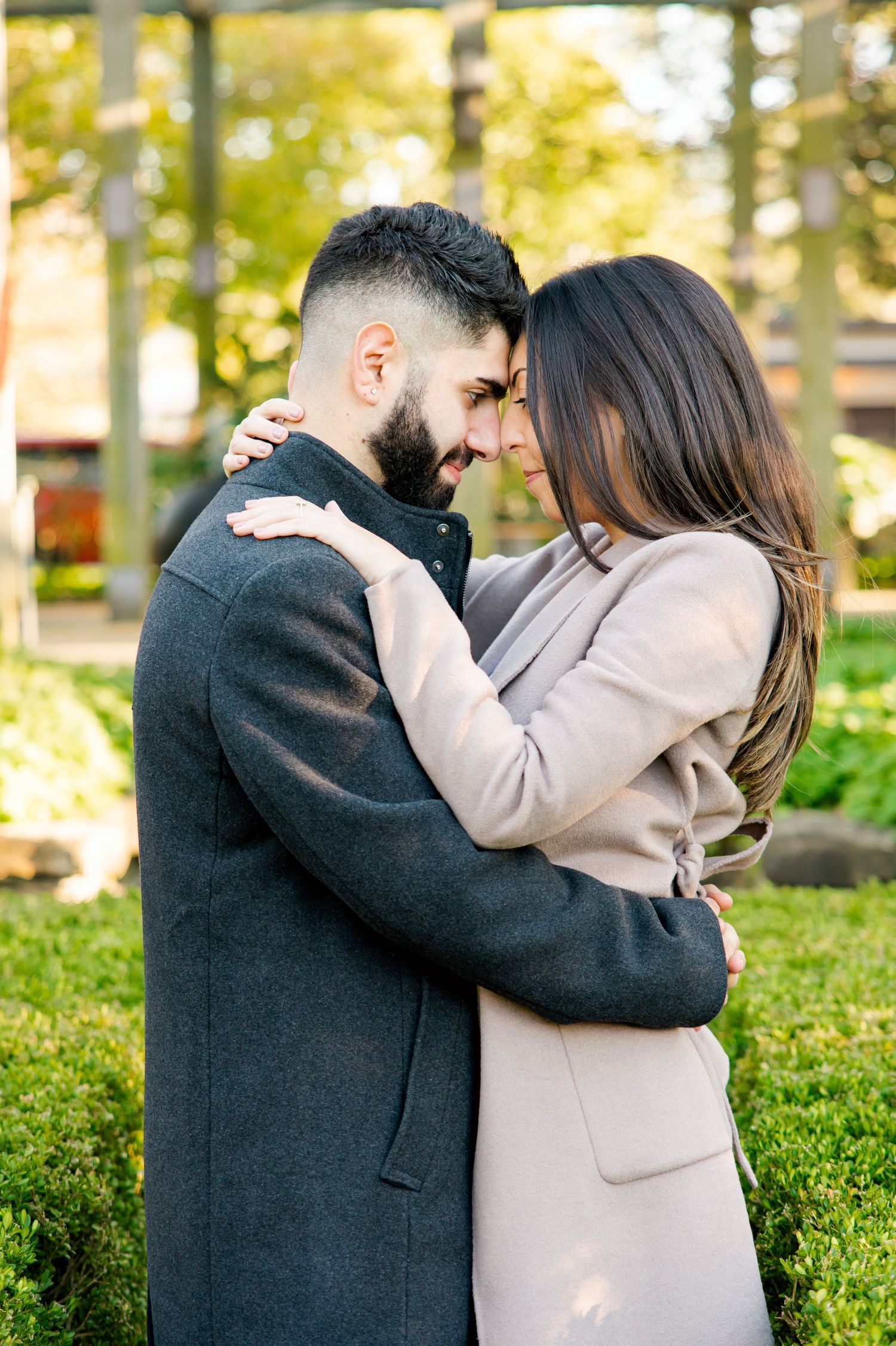 Intimate portrait of couple embracing in a sunlit garden wearing long coats.
