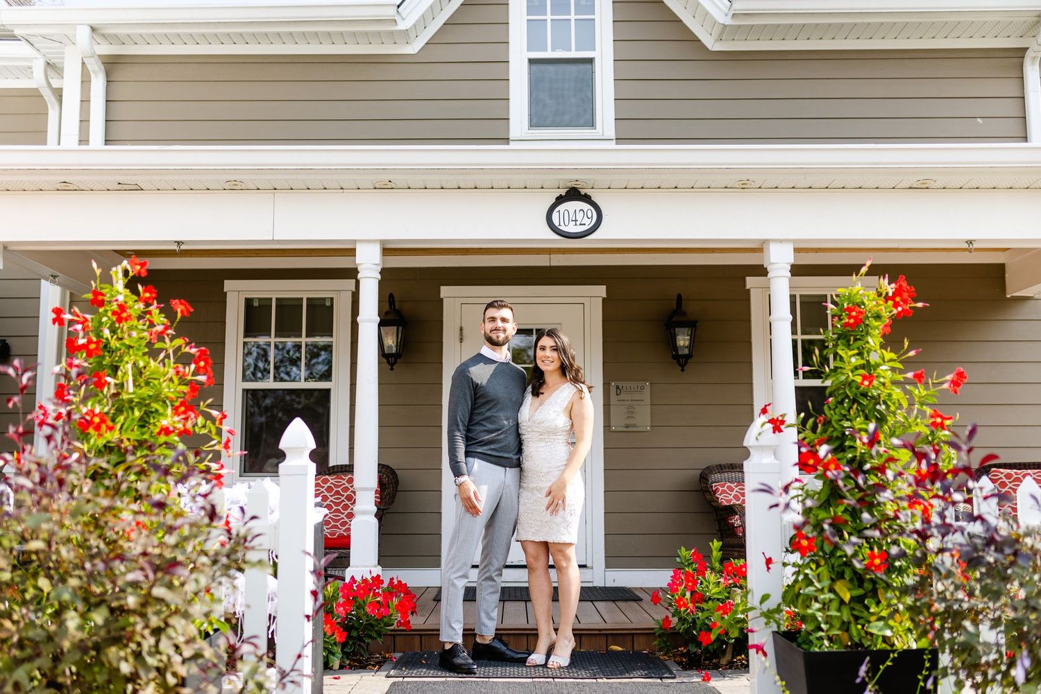 Two people standing on front porch decorated with vibrant flowers and red planters.