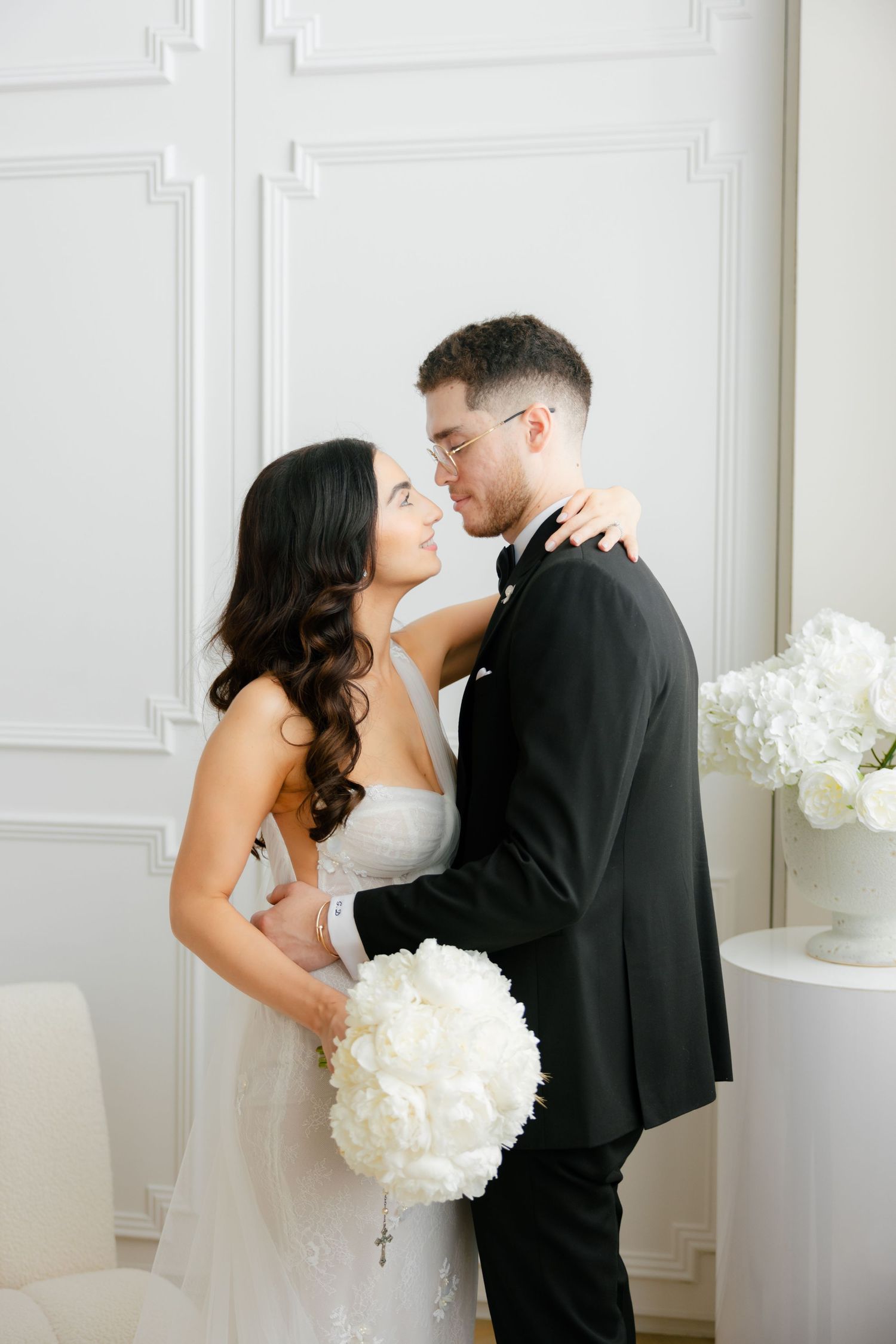 A romantic wedding portrait of a couple embracing in a bright white room with a beautiful floral bouquet.