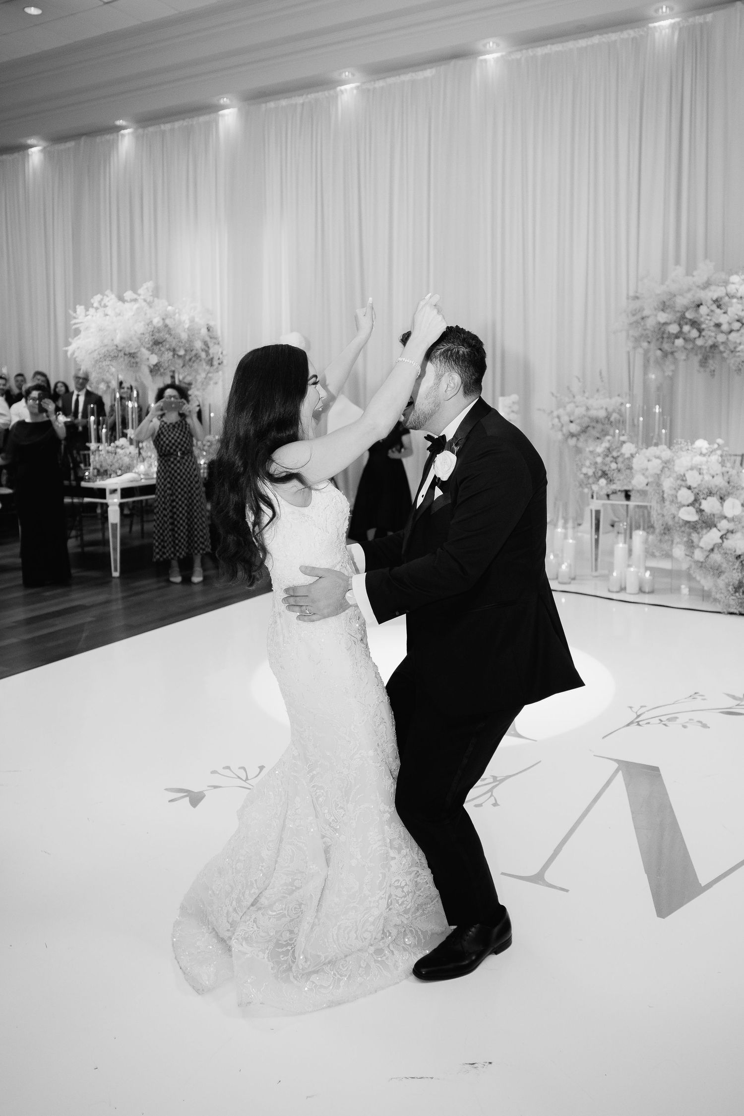 A black and white photo captures a couple dancing energetically on a white dance floor at their wedding.