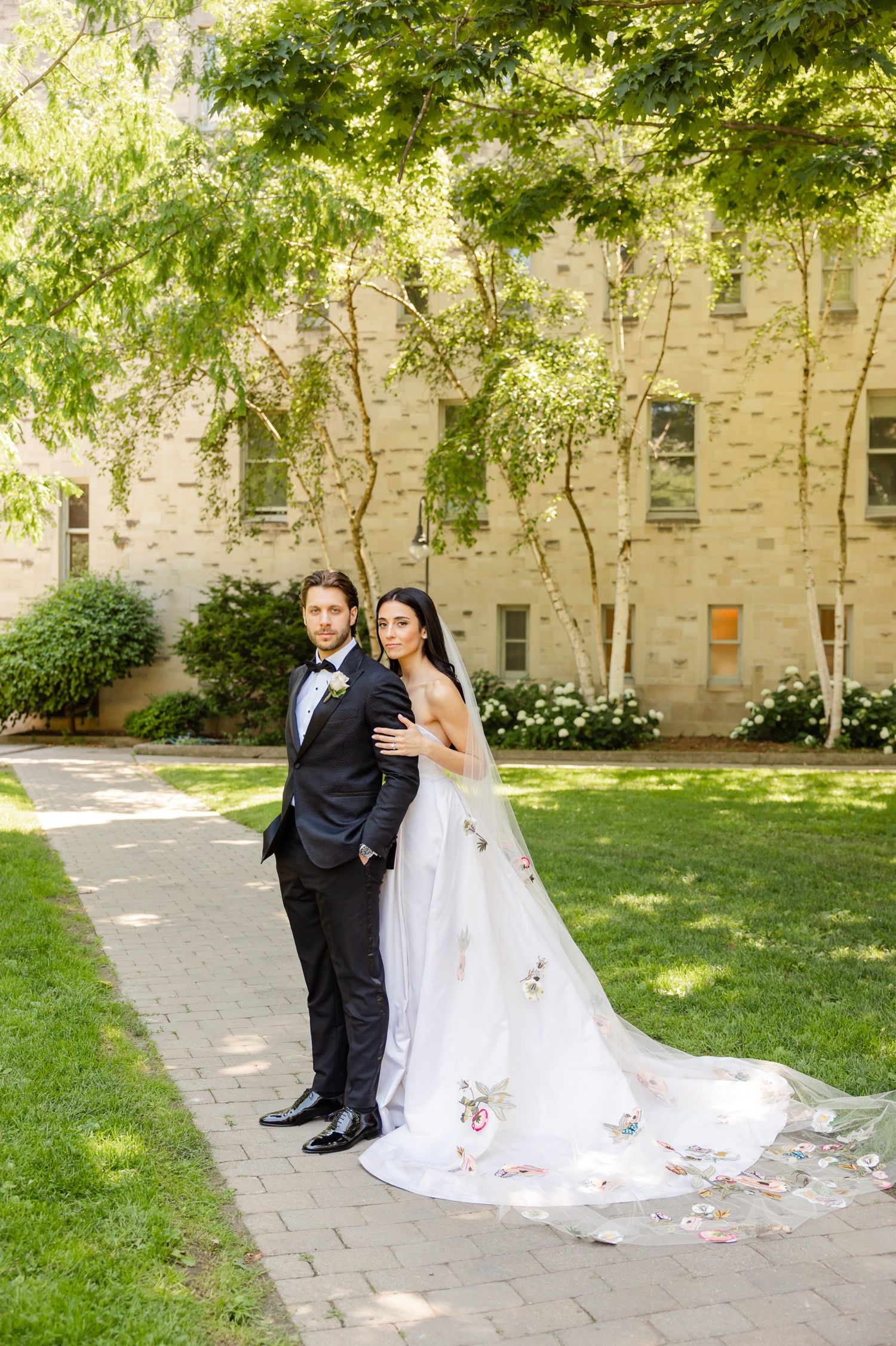 A couple poses on a brick path with a beautiful stone building and trees in the background.