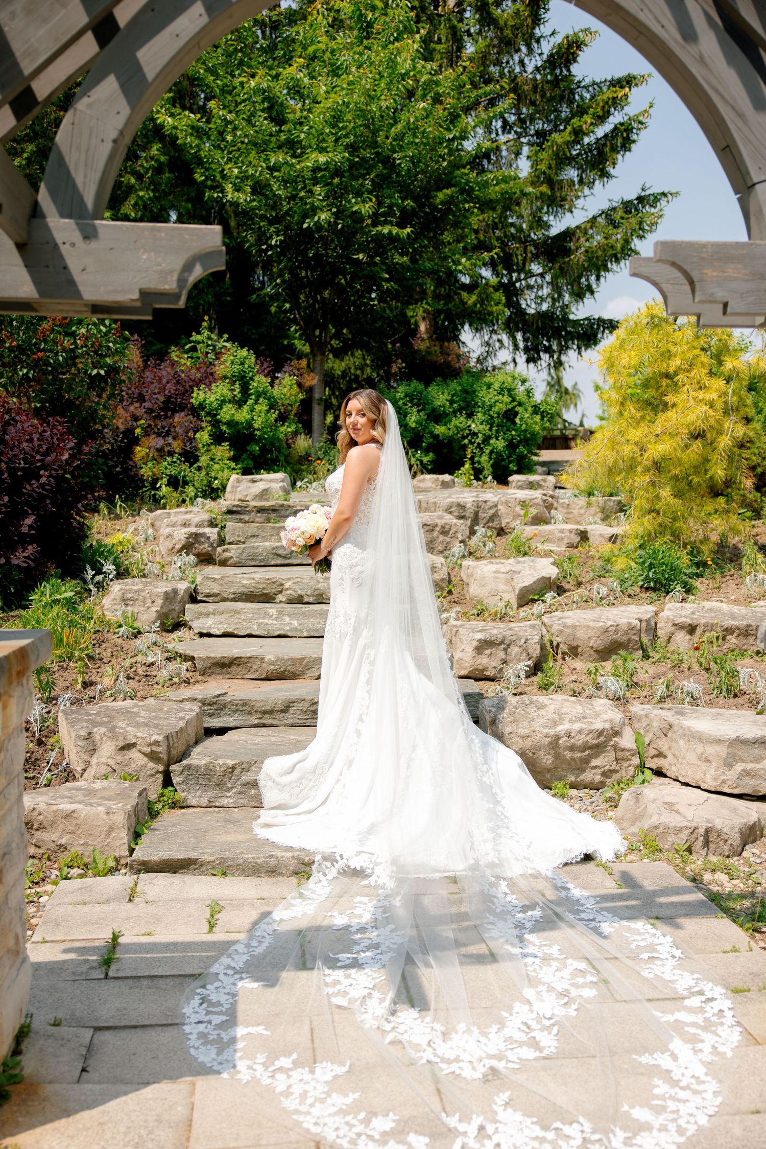 A bride in an elegant white gown with a dramatic cathedral veil poses on stone garden steps under an arch.
