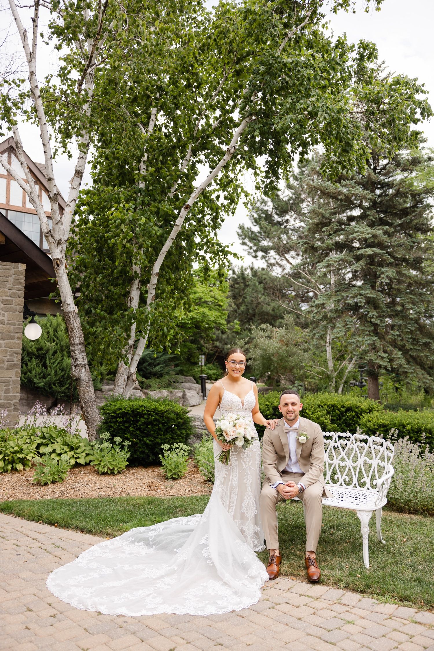 A wedding couple poses in a garden setting with a white wicker chair and flowing lace dress train on brick pavers.