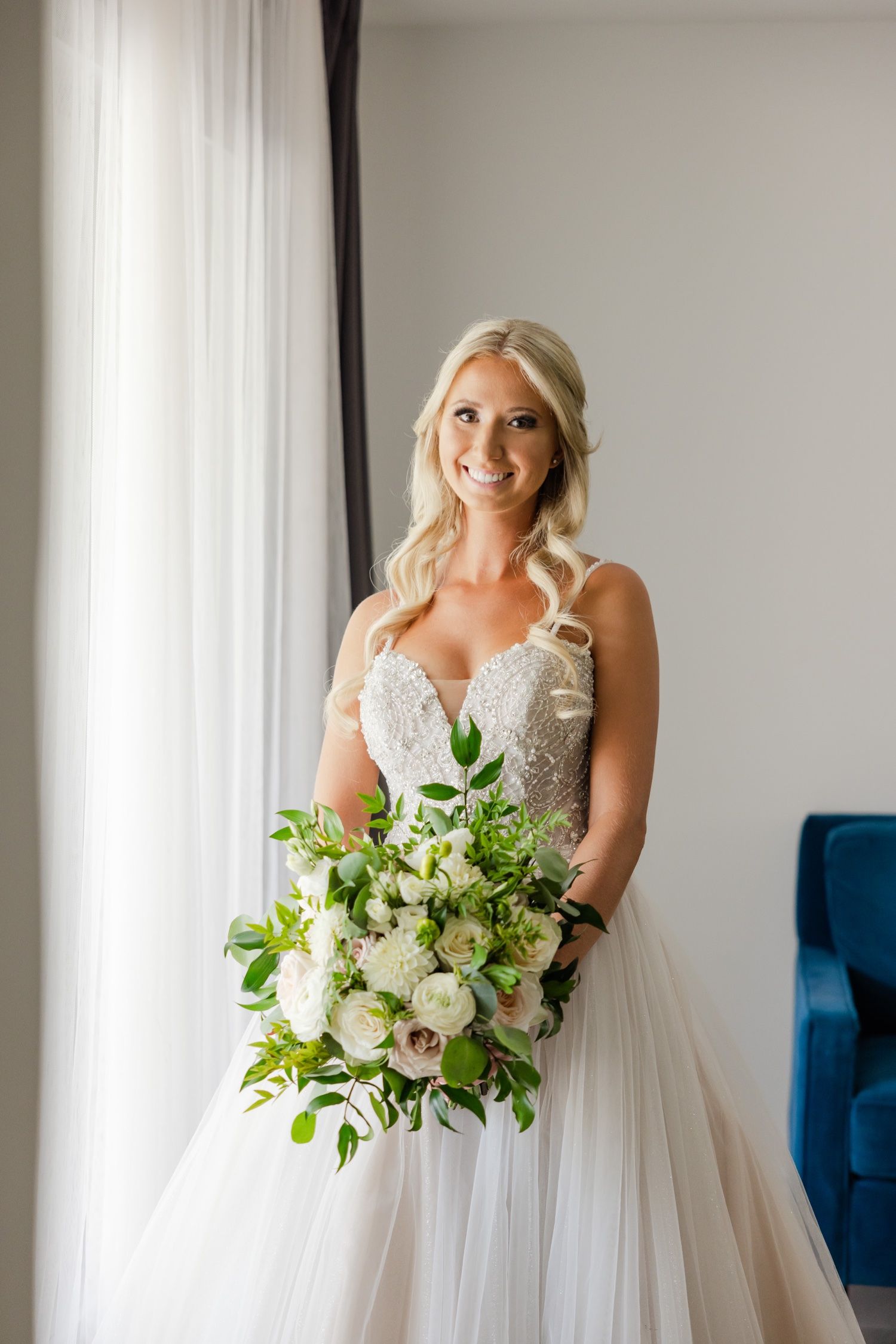 Bride in detailed sweetheart neckline gown holds a fresh white and green bouquet by the window.