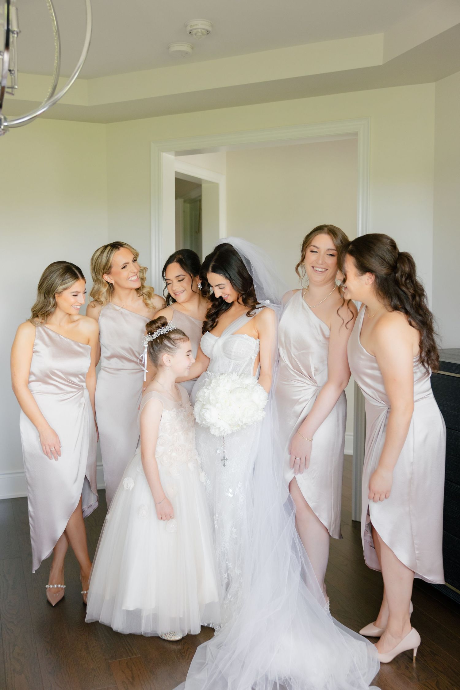 A bridal party wearing light pink bridesmaid dresses and a white wedding gown shares a candid moment indoors.