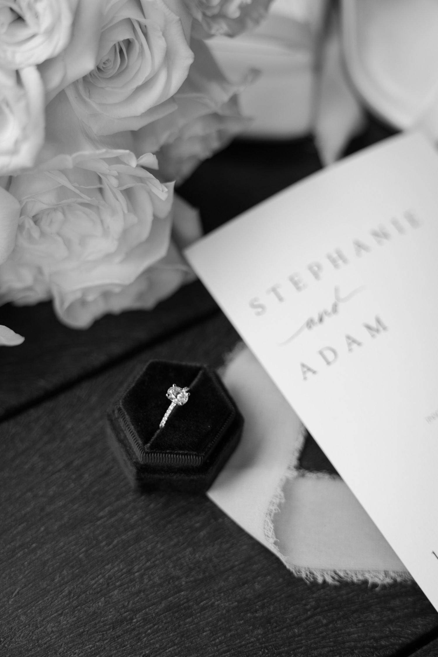 Elegant white roses next to a wedding invitation and engagement ring in a black velvet box.