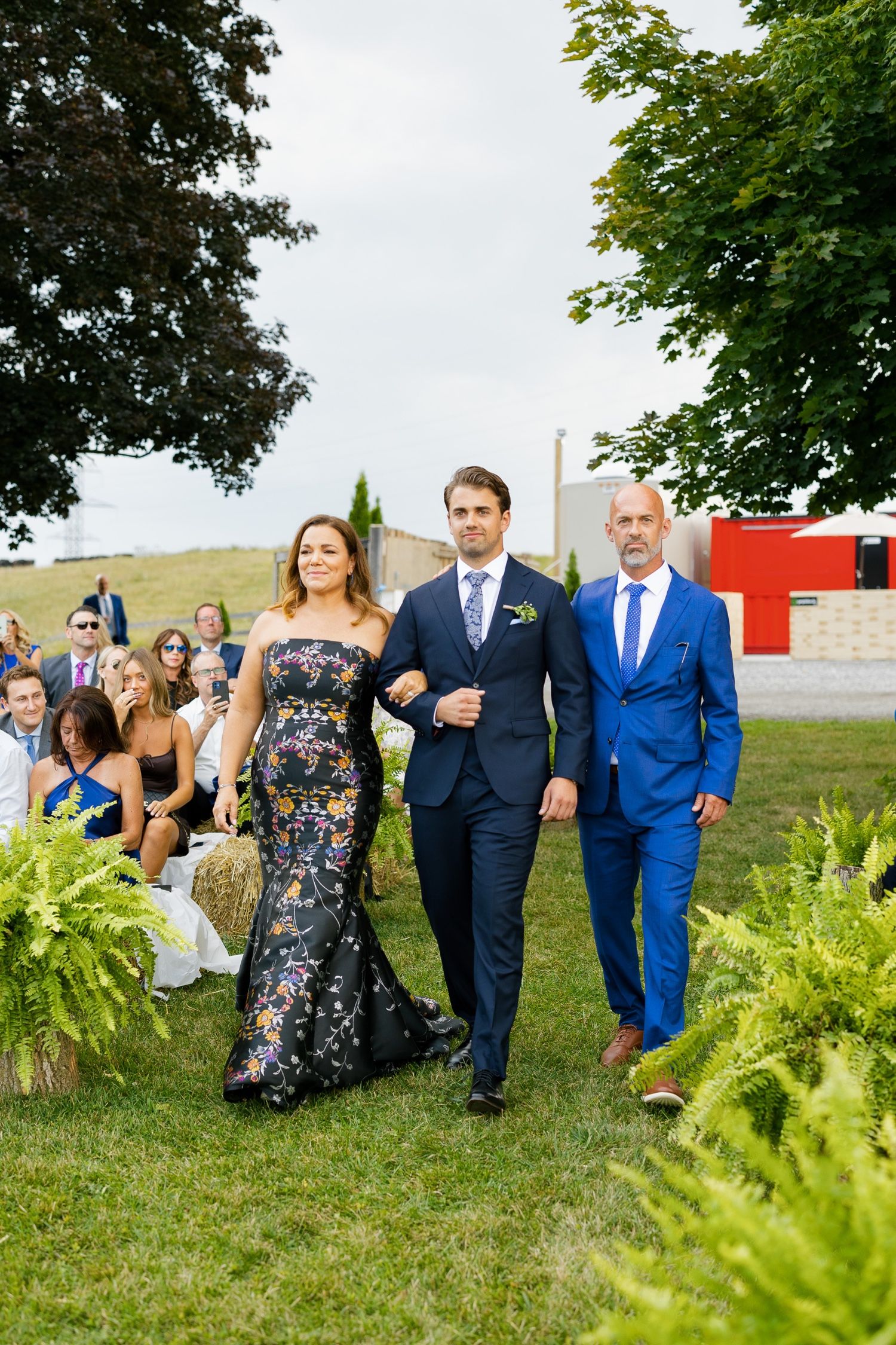 Wedding party members walk down grassy aisle lined with ferns during outdoor ceremony.