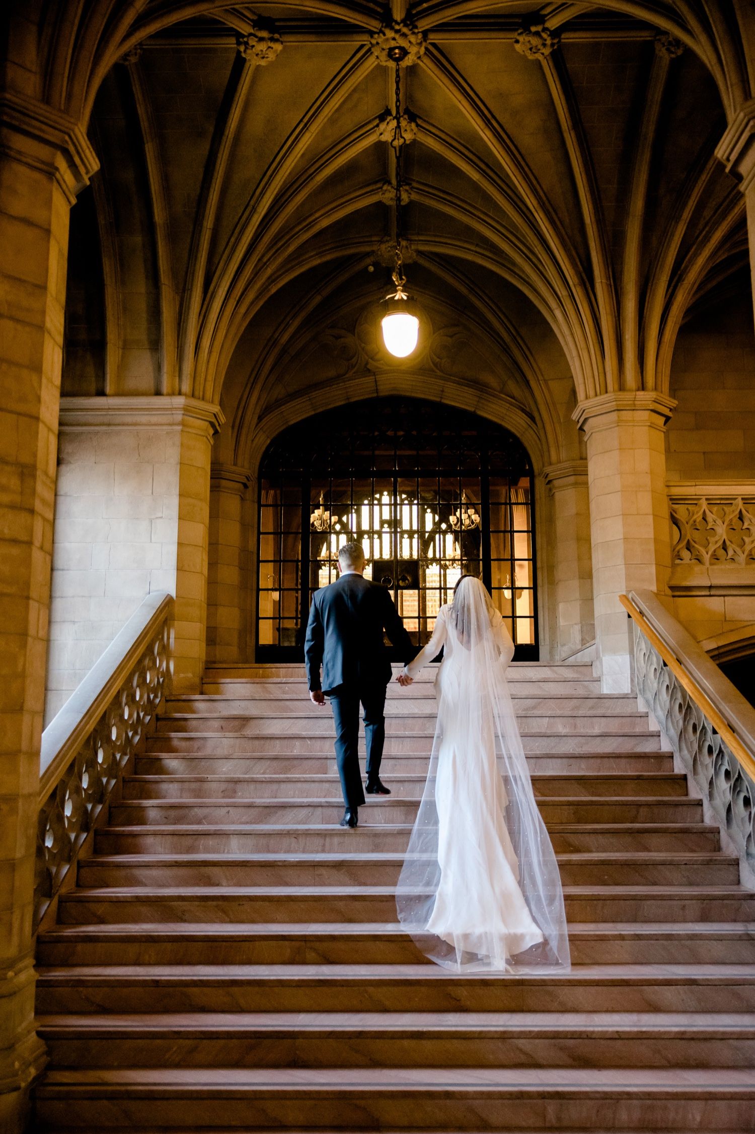 A couple ascends an elegant Gothic staircase with vaulted ceilings and ornate architecture.