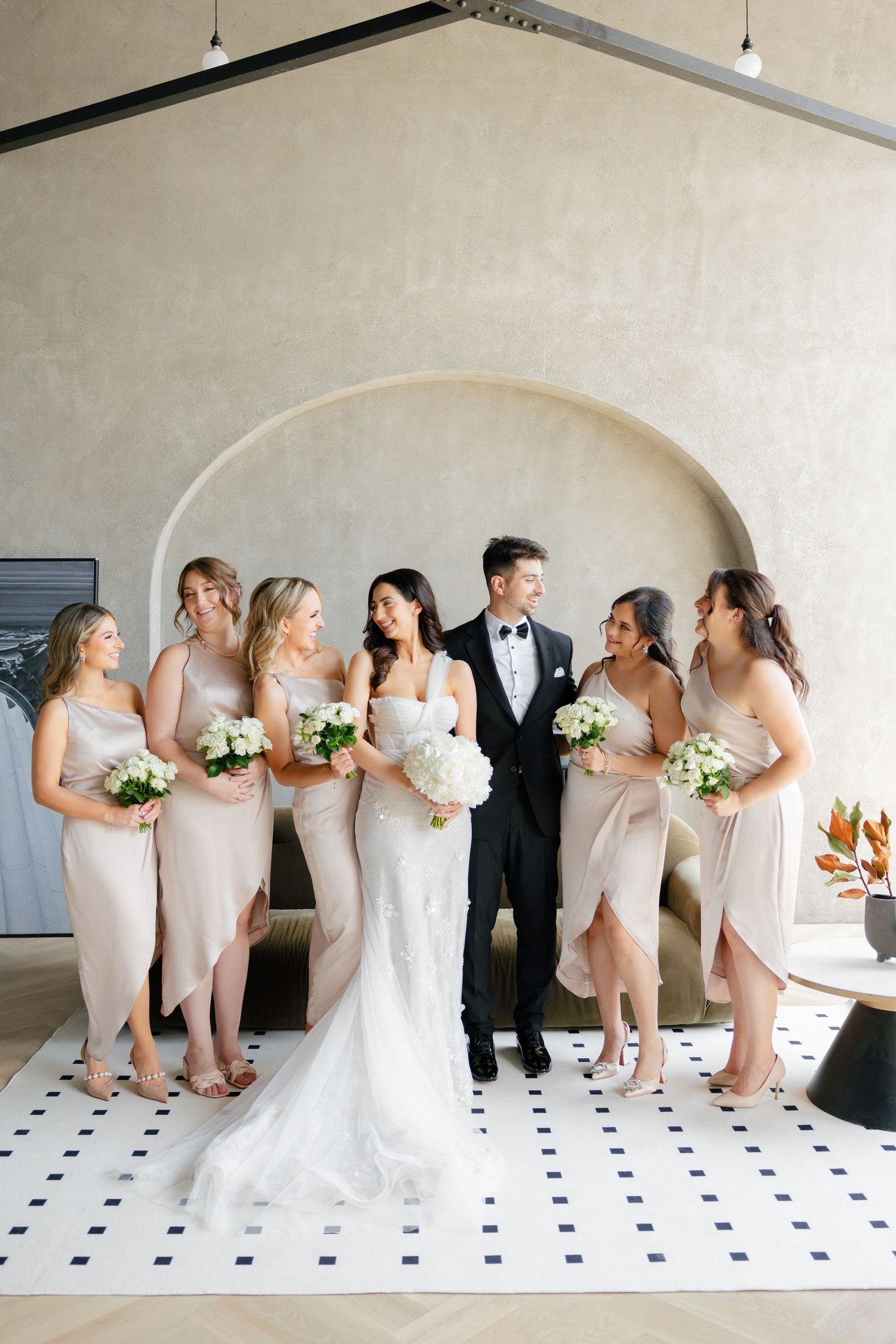 Wedding party laughs together while posing for formal photos under archway.