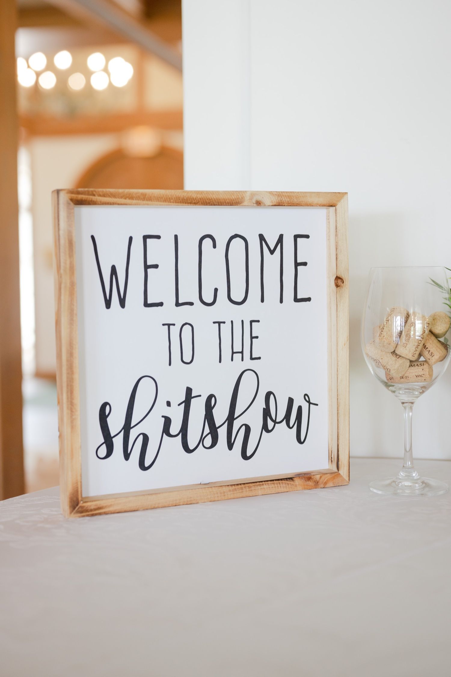 Rustic wooden framed welcome sign reading 'Welcome to the Shitshow' next to wine glass.