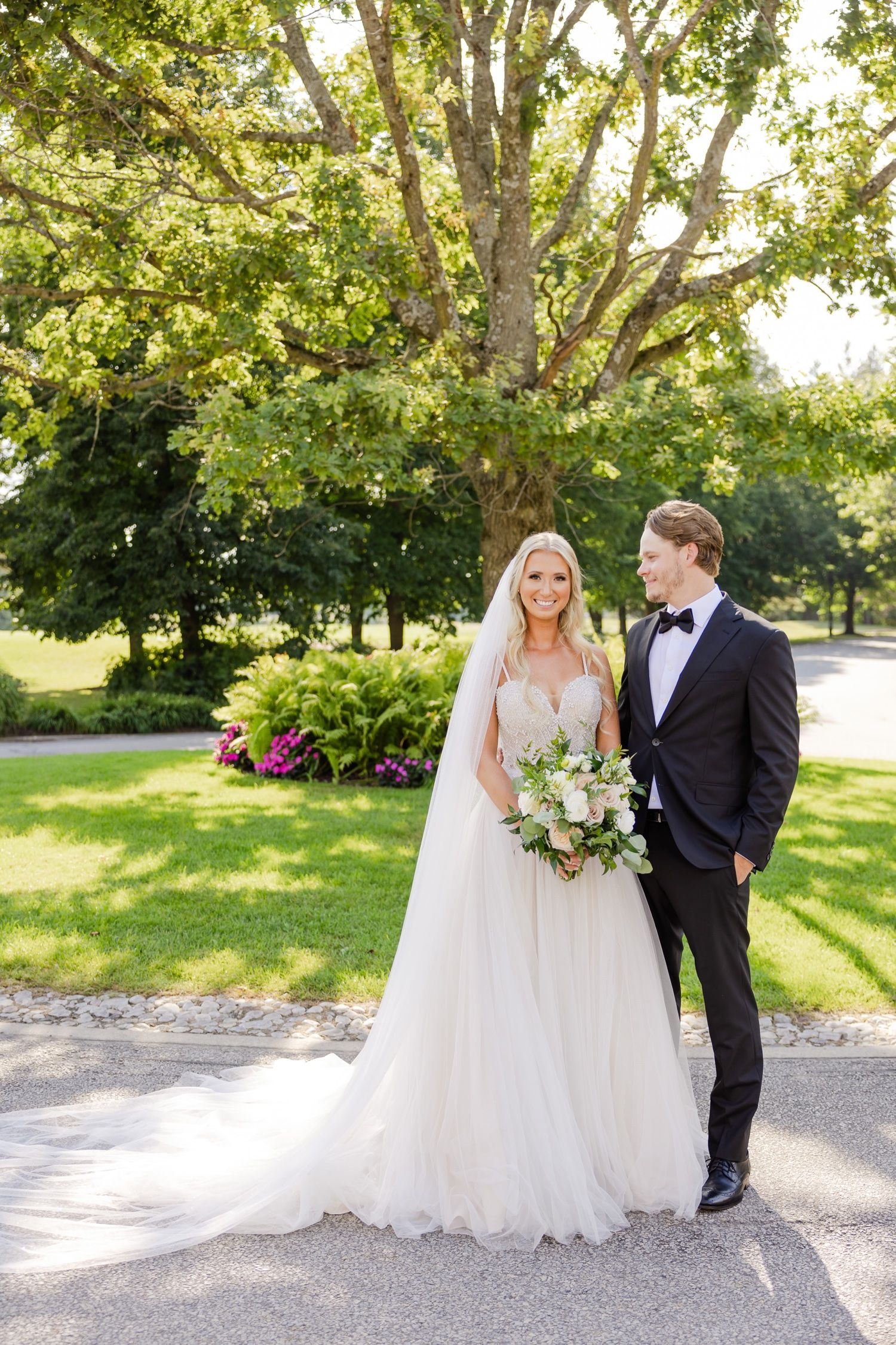 Elegant wedding couple standing under a large tree in a manicured garden setting.