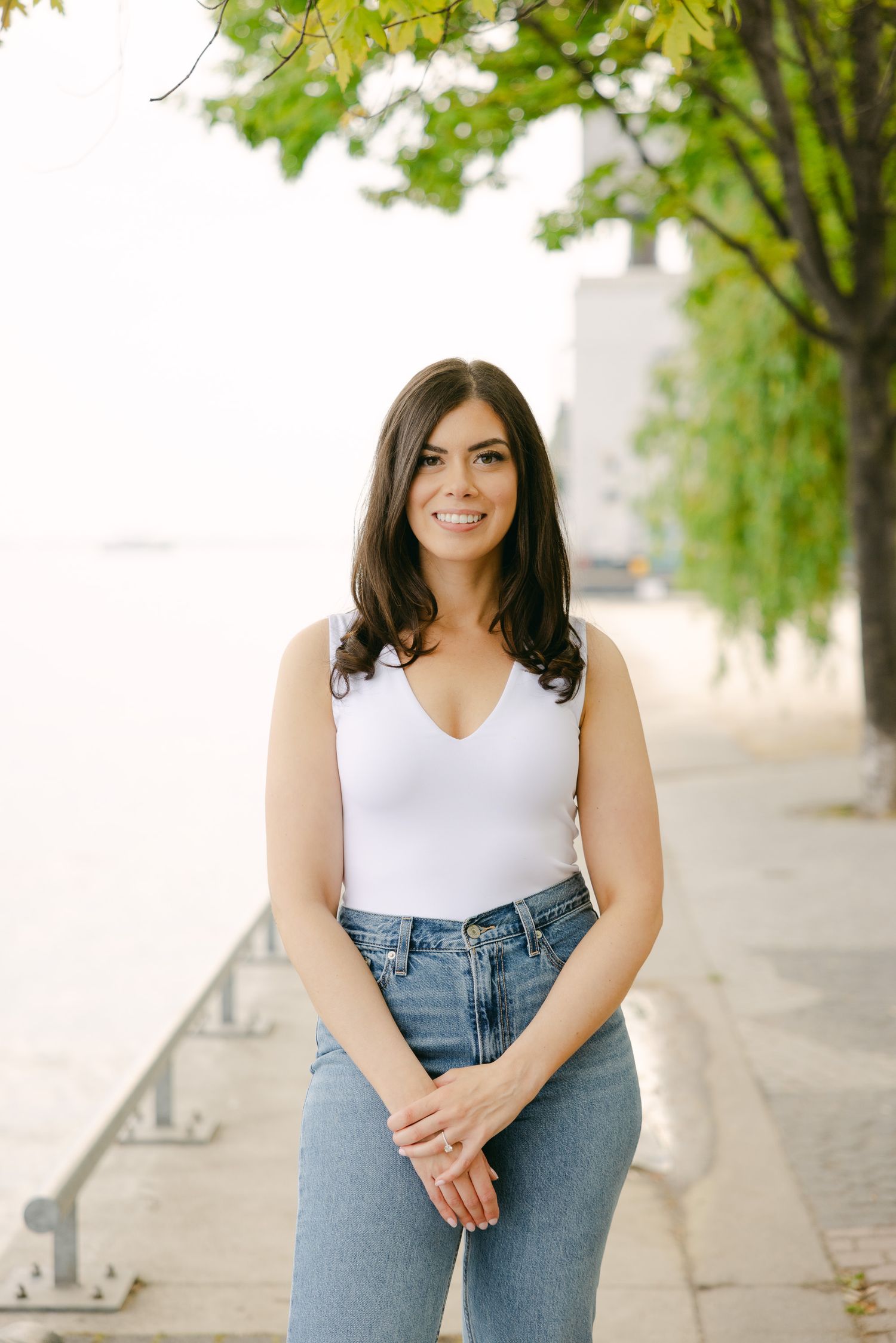 Portrait of person in white sleeveless top and jeans standing on waterfront path.