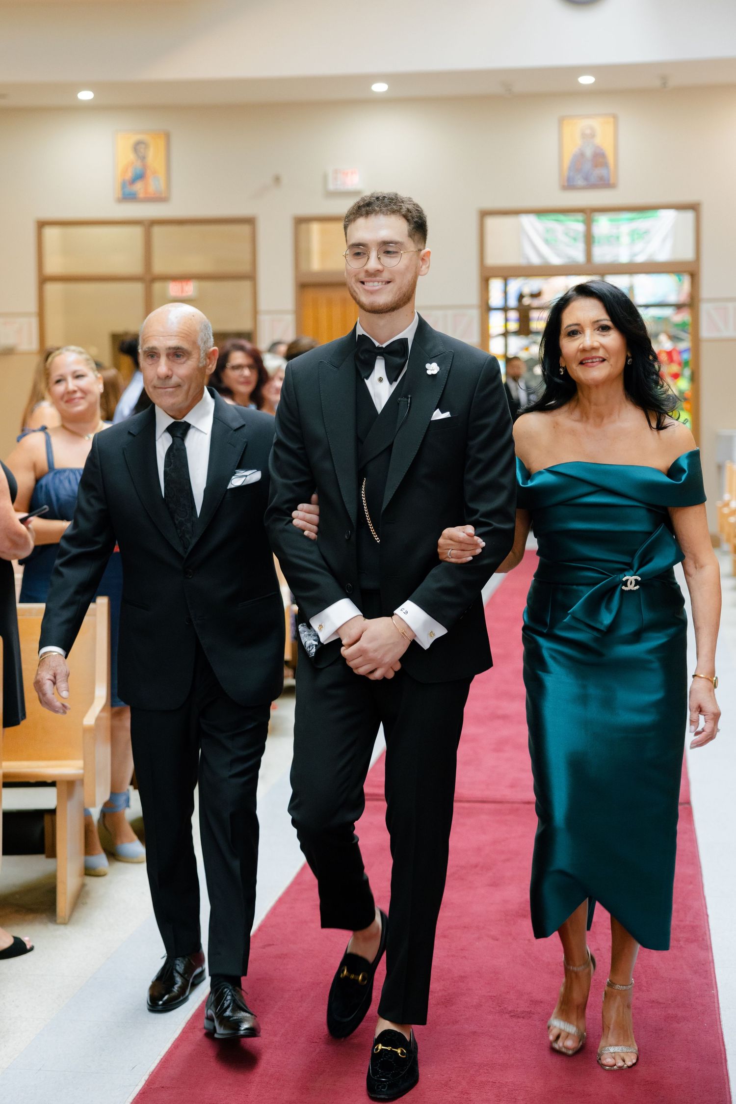 Wedding party members walk arm-in-arm down church aisle with guests seated in pews.