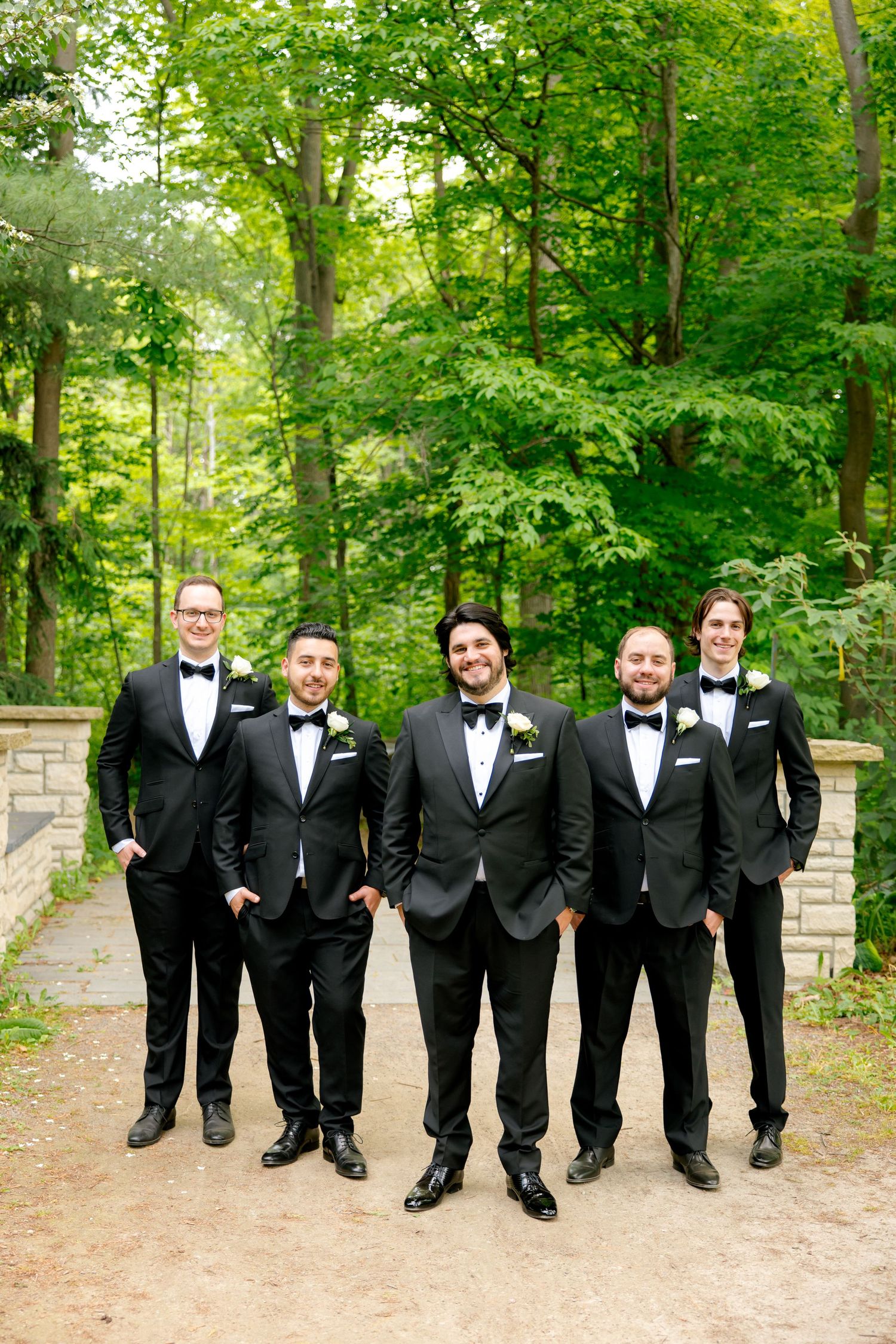 Five groomsmen in classic black tuxedos standing on garden path surrounded by green foliage.