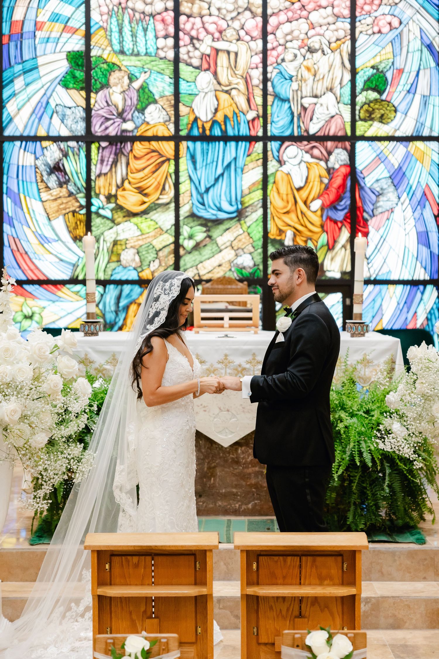 Couple exchanging vows in front of a vibrant stained glass window with white floral arrangements and ferns.