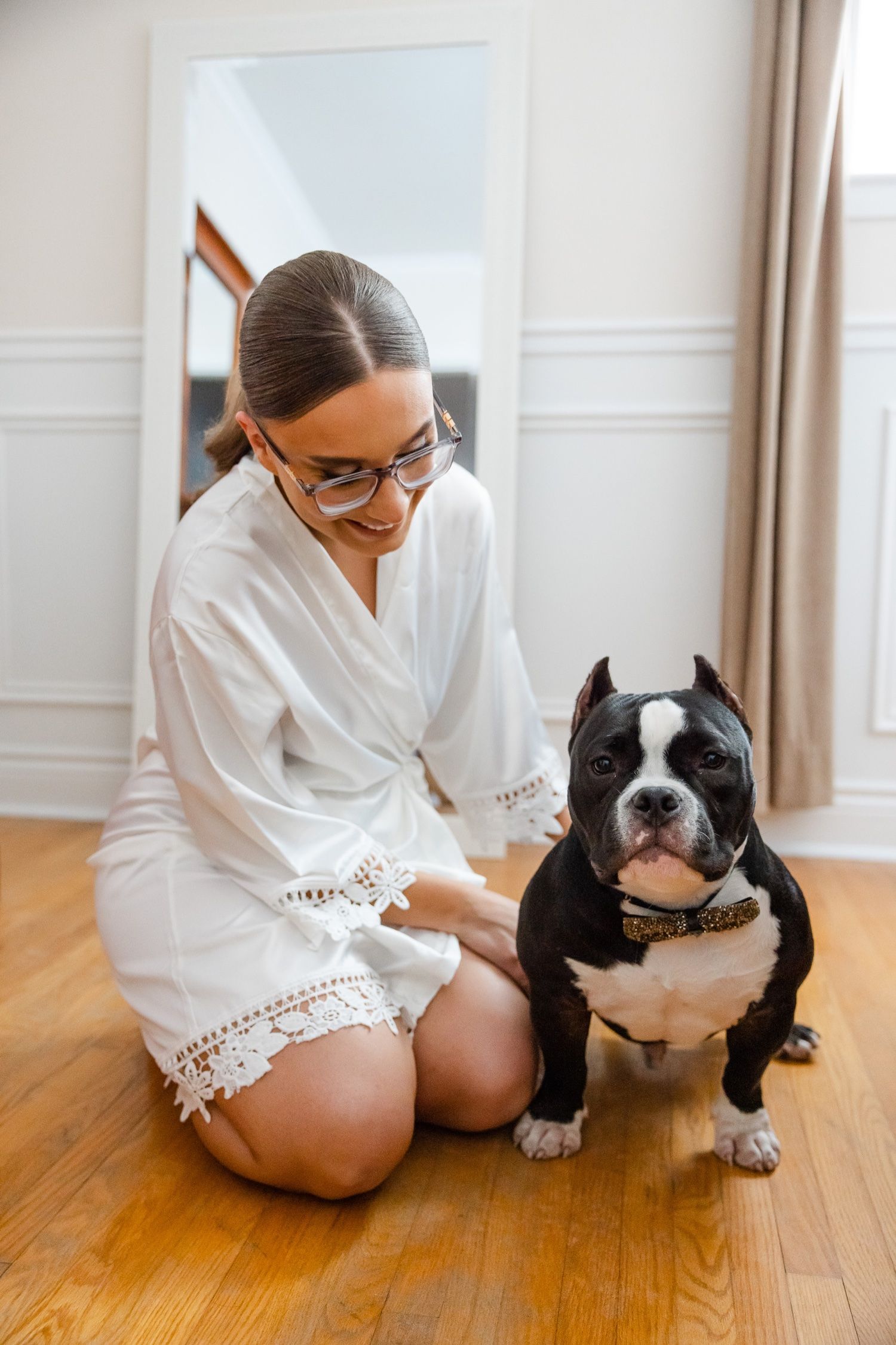 Person in white robe kneeling beside Boston Terrier on hardwood floor.
