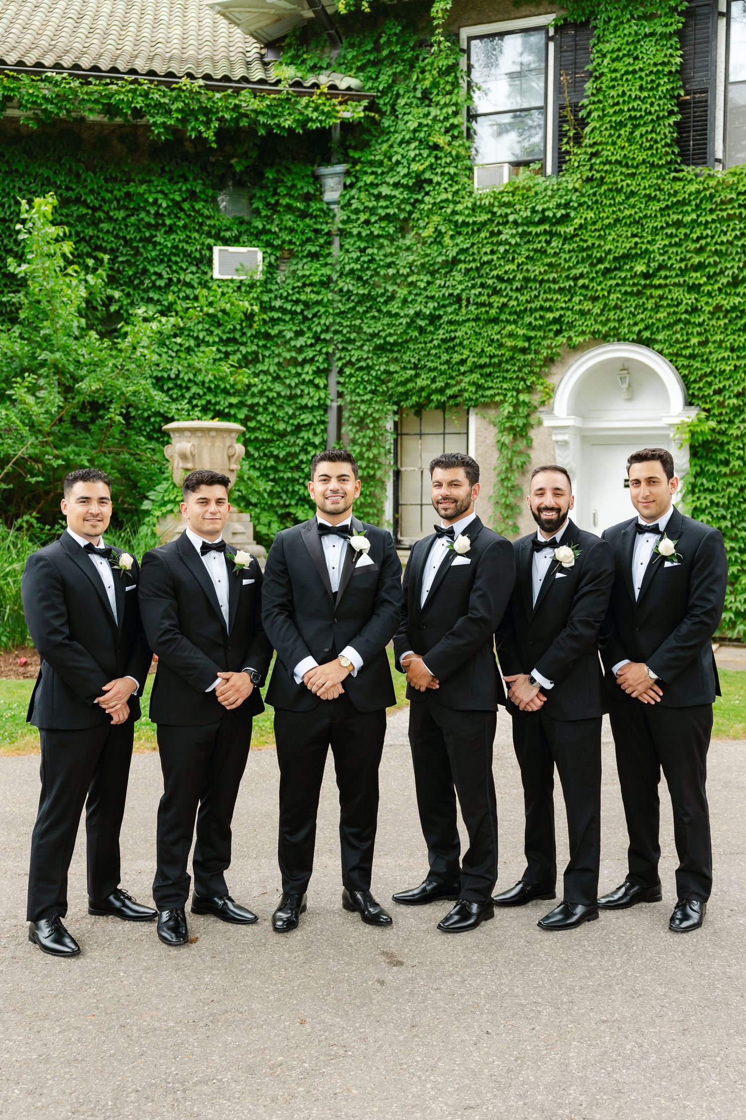 Groomsmen in matching black tuxedos pose casually in front of ivy-covered building facade.