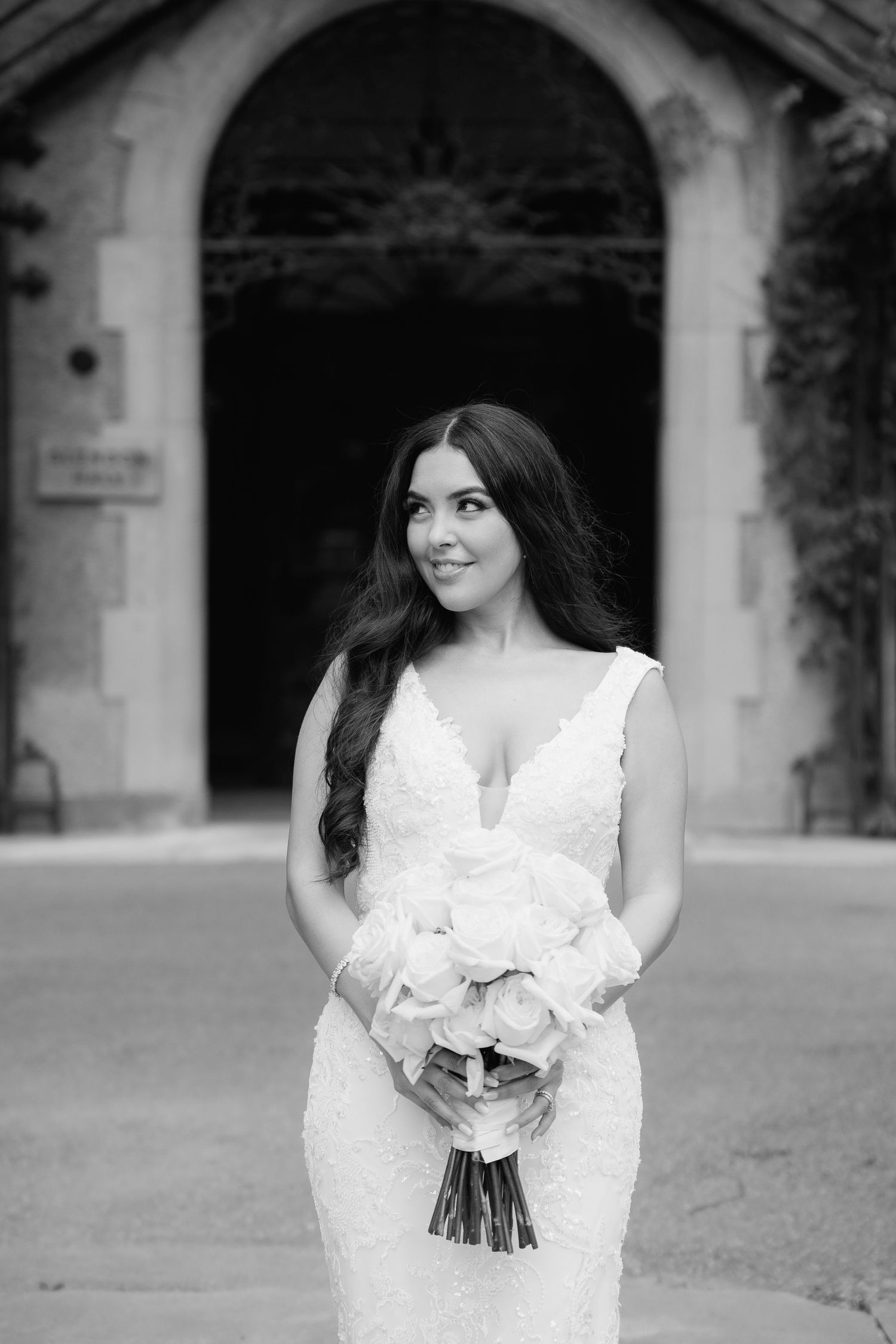 Black and white portrait of bride holding bouquet under arched stone entrance with trailing ivy.