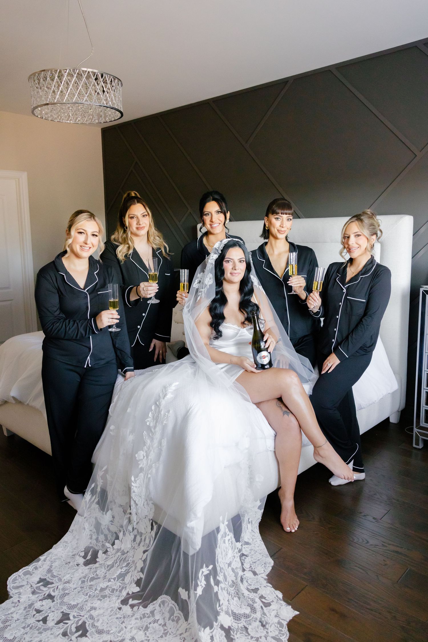 Wedding party poses on bed with champagne, bridesmaids in black and bride in white lace gown.