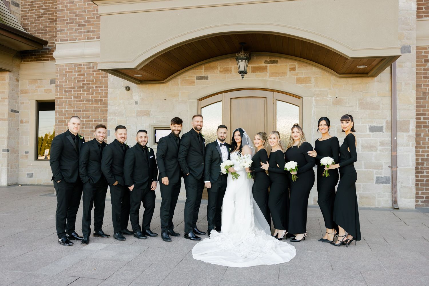 Wedding party in formal black attire and white bridal gown poses together on stone building steps.