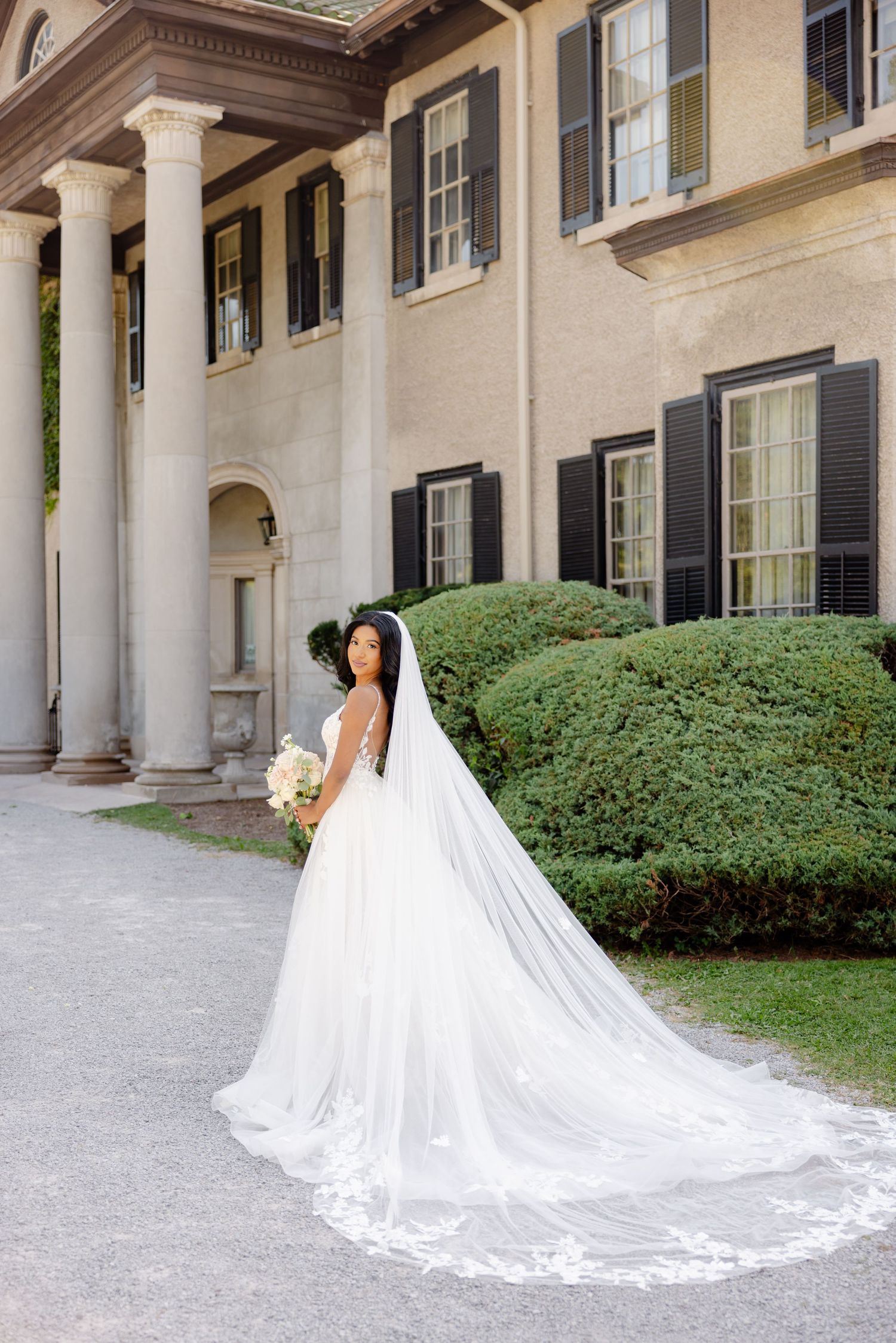 An elegant bride in a flowing white dress and veil stands before a classical building with ornate columns.