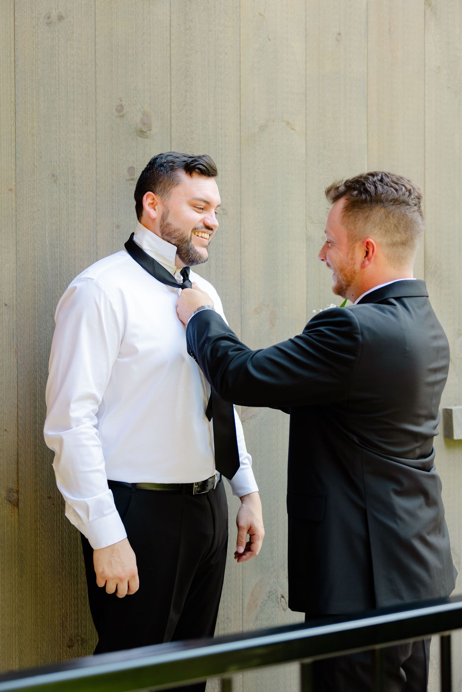 Groomsman helps adjust another man's tie before wedding ceremony against wooden wall.