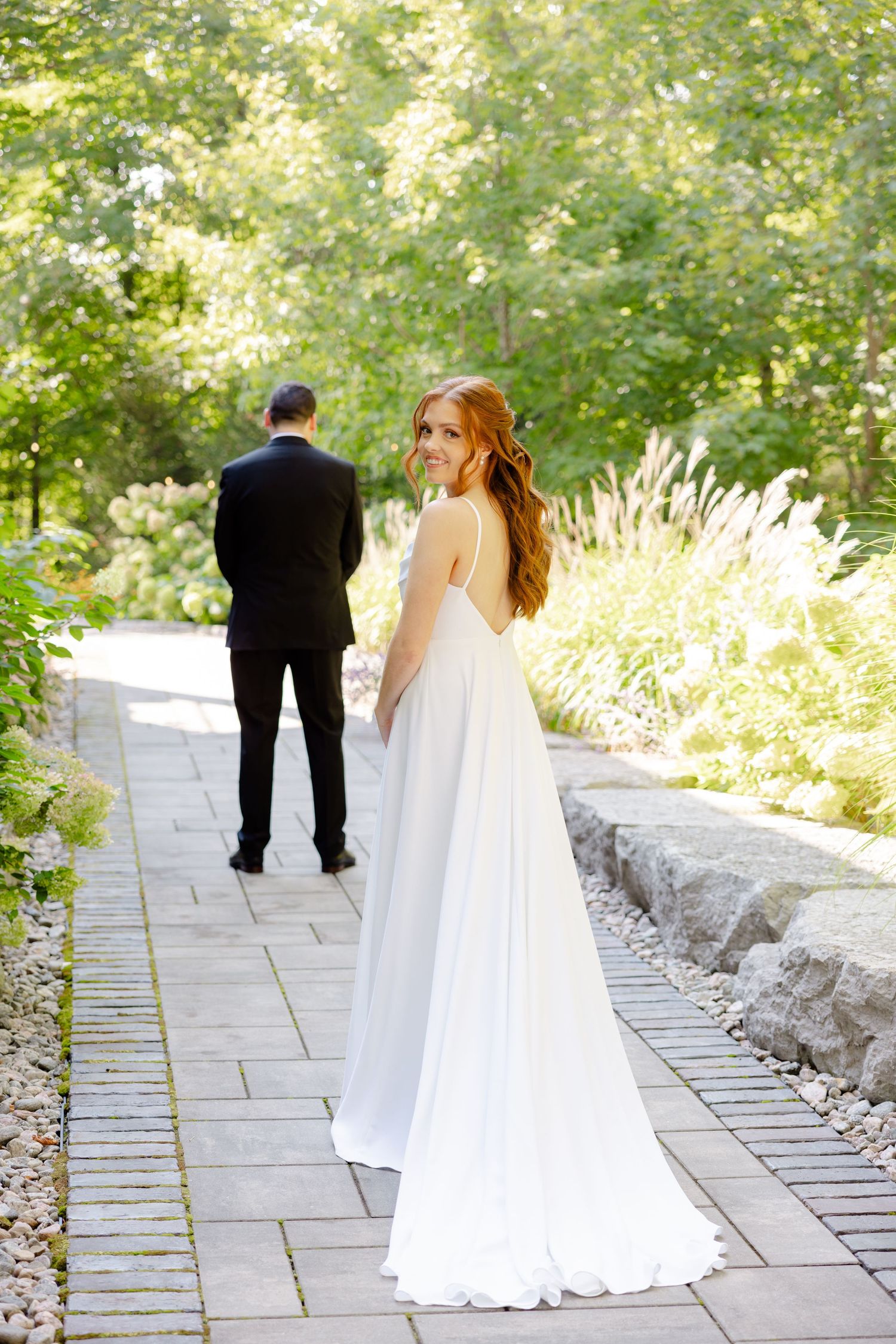 Bride in flowing white gown looks back while groom faces away on garden pathway.
