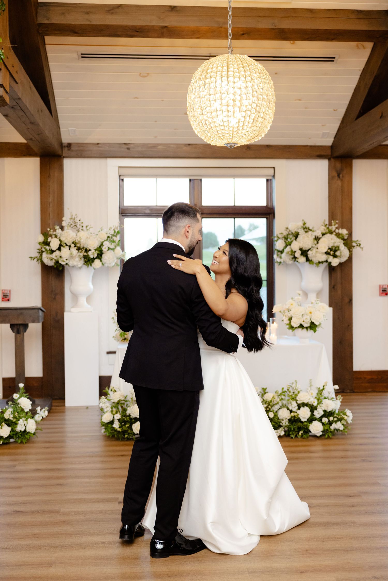 A couple embraces while dancing in a rustic reception venue decorated with white flowers and wooden accents.