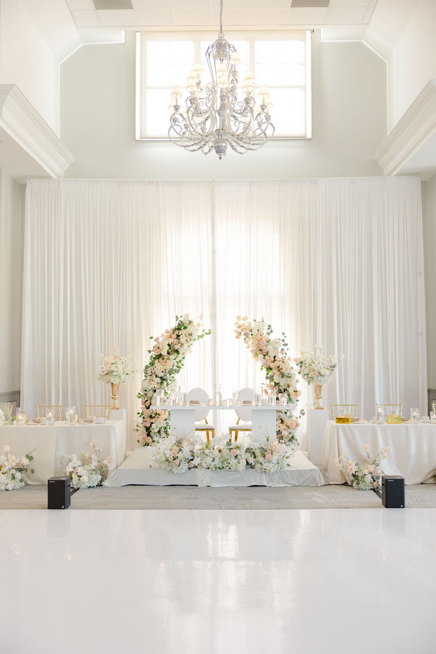White wedding ceremony space with floral arch and crystal chandelier in elegant ballroom.