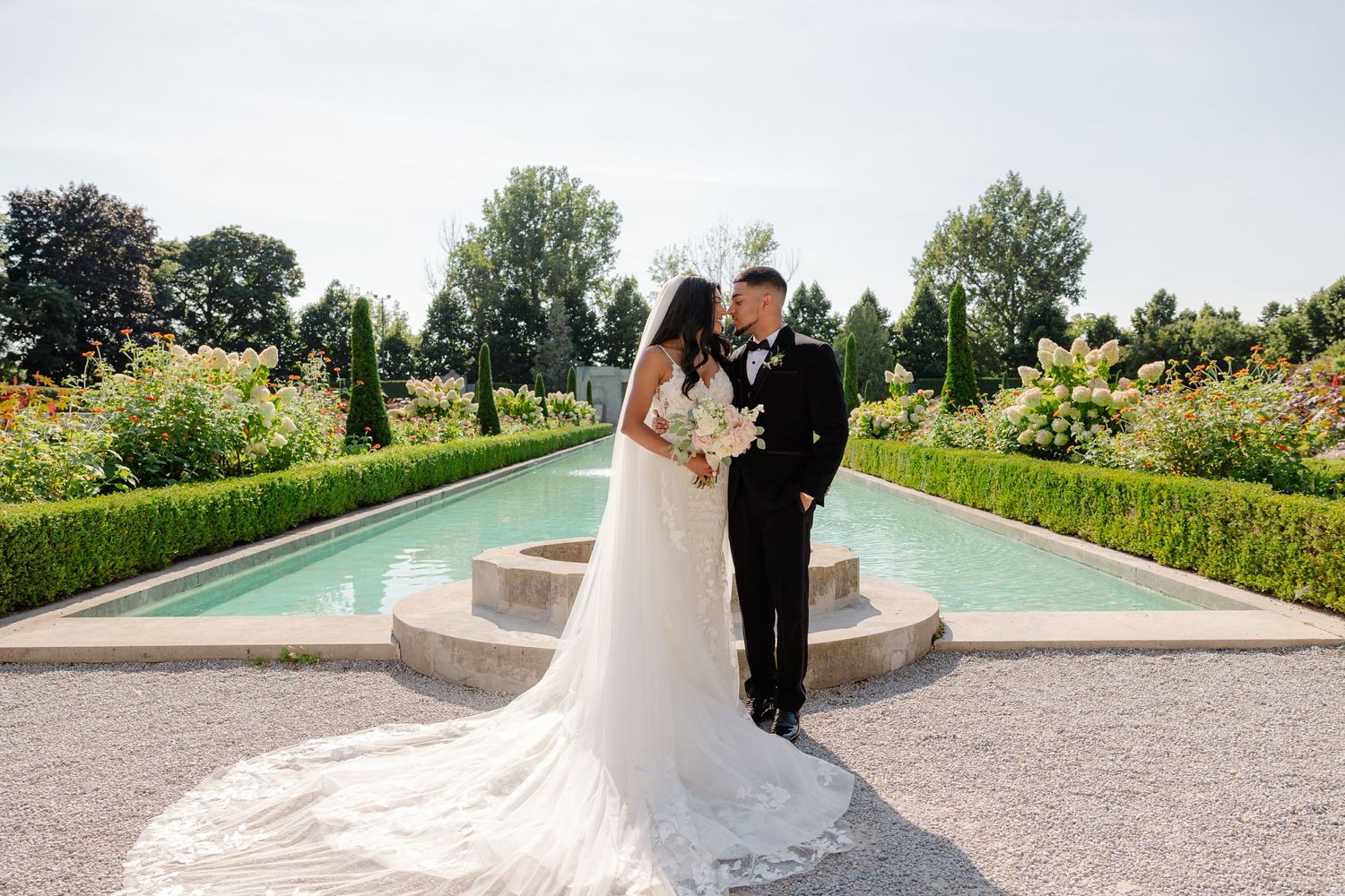 Newlyweds pose together in front of an elegant garden fountain with flowing water.