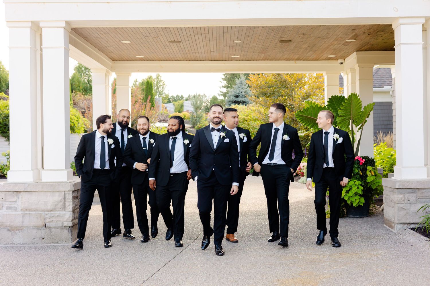 A group of groomsmen in black tuxedos walking together under a covered outdoor walkway.