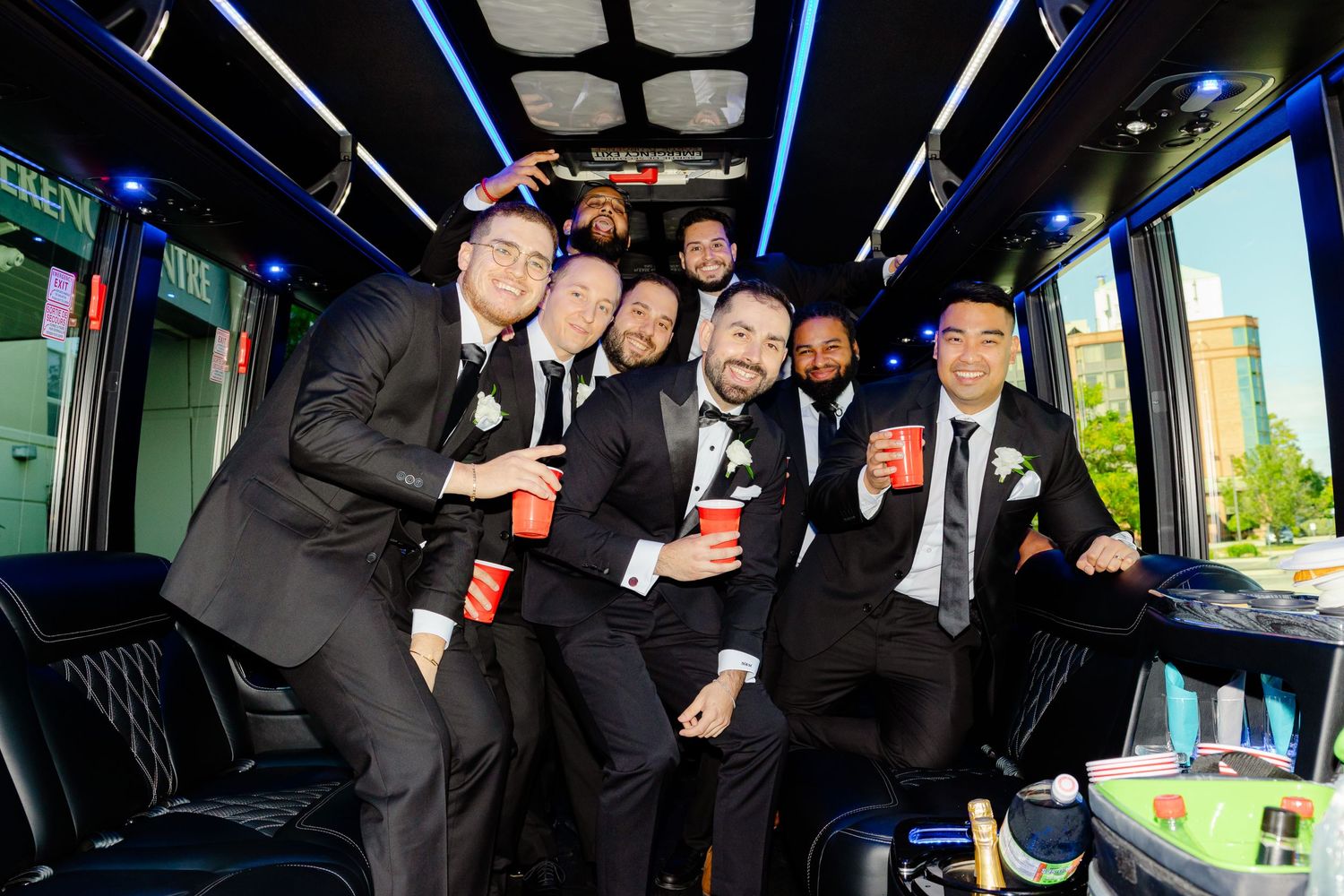 Group of formally dressed people celebrating with drinks inside a party bus with blue lighting.