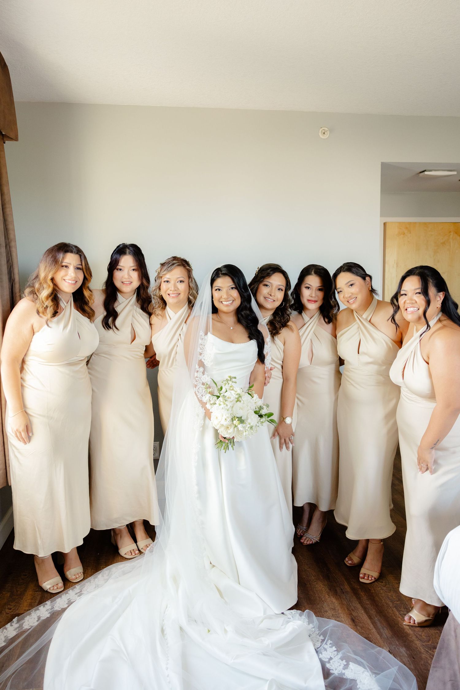 Wedding party poses together in neutral-toned dresses against a light wall.