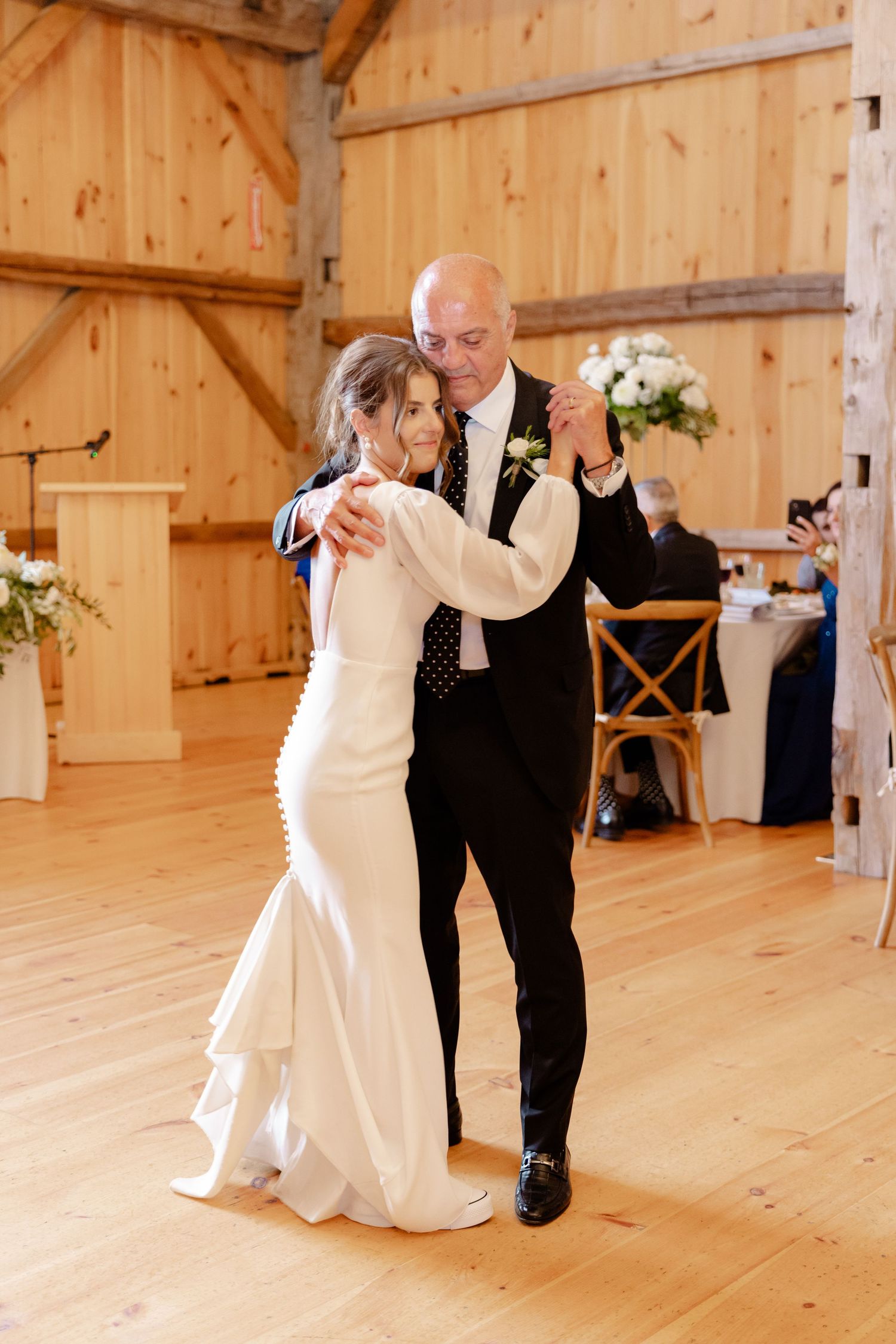 Couple dancing together on wooden floor of rustic barn venue with floral decorations.