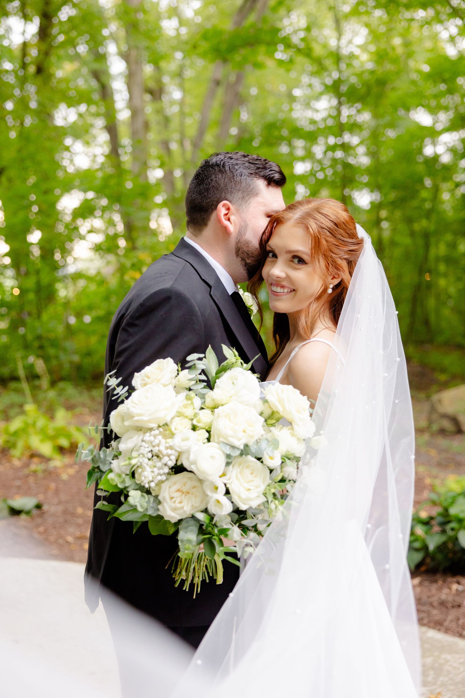 A tender wedding moment captured outdoors with white roses and flowing veil against green foliage.