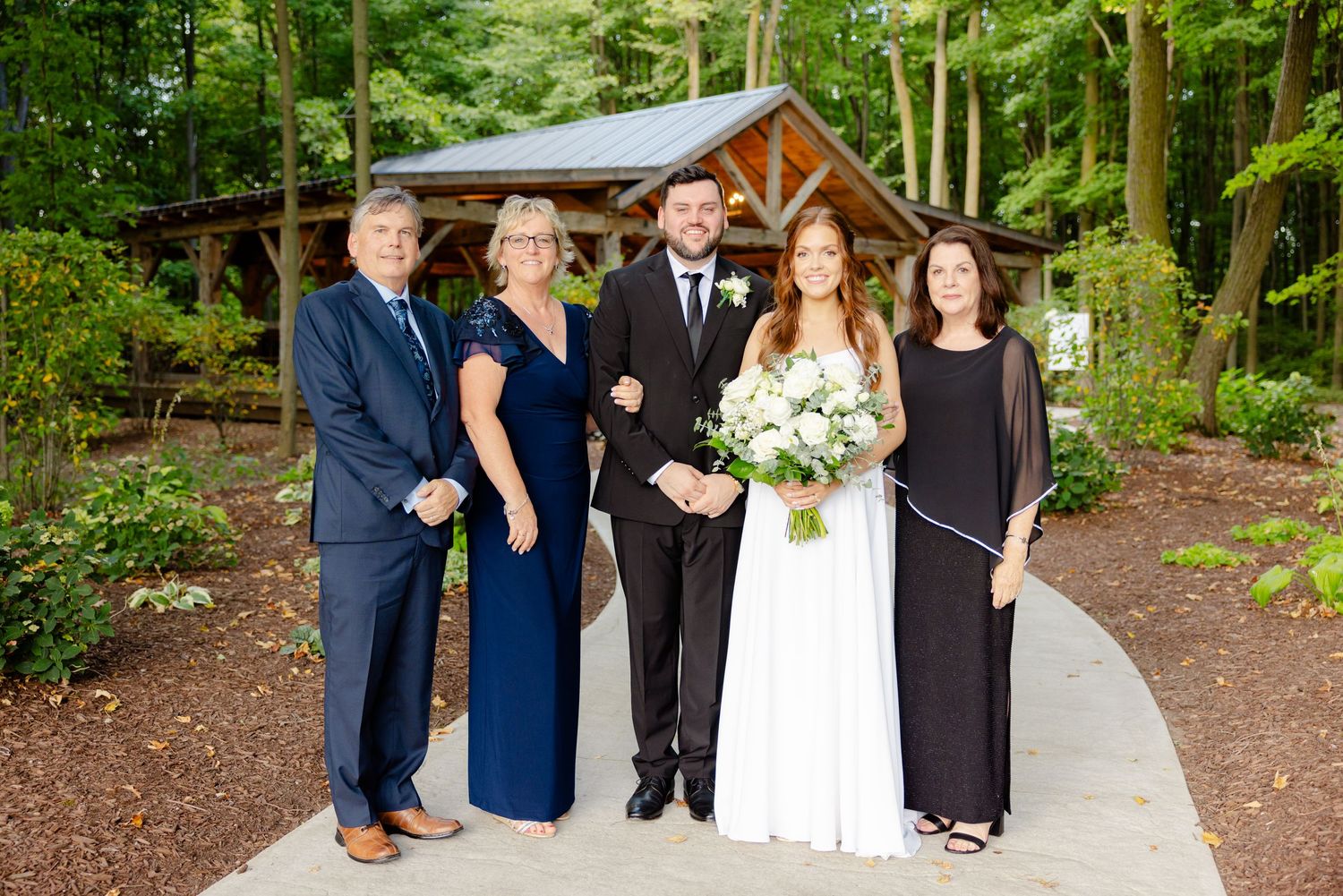 Wedding party members pose in formal attire on a paved path with a rustic wooden pavilion backdrop.