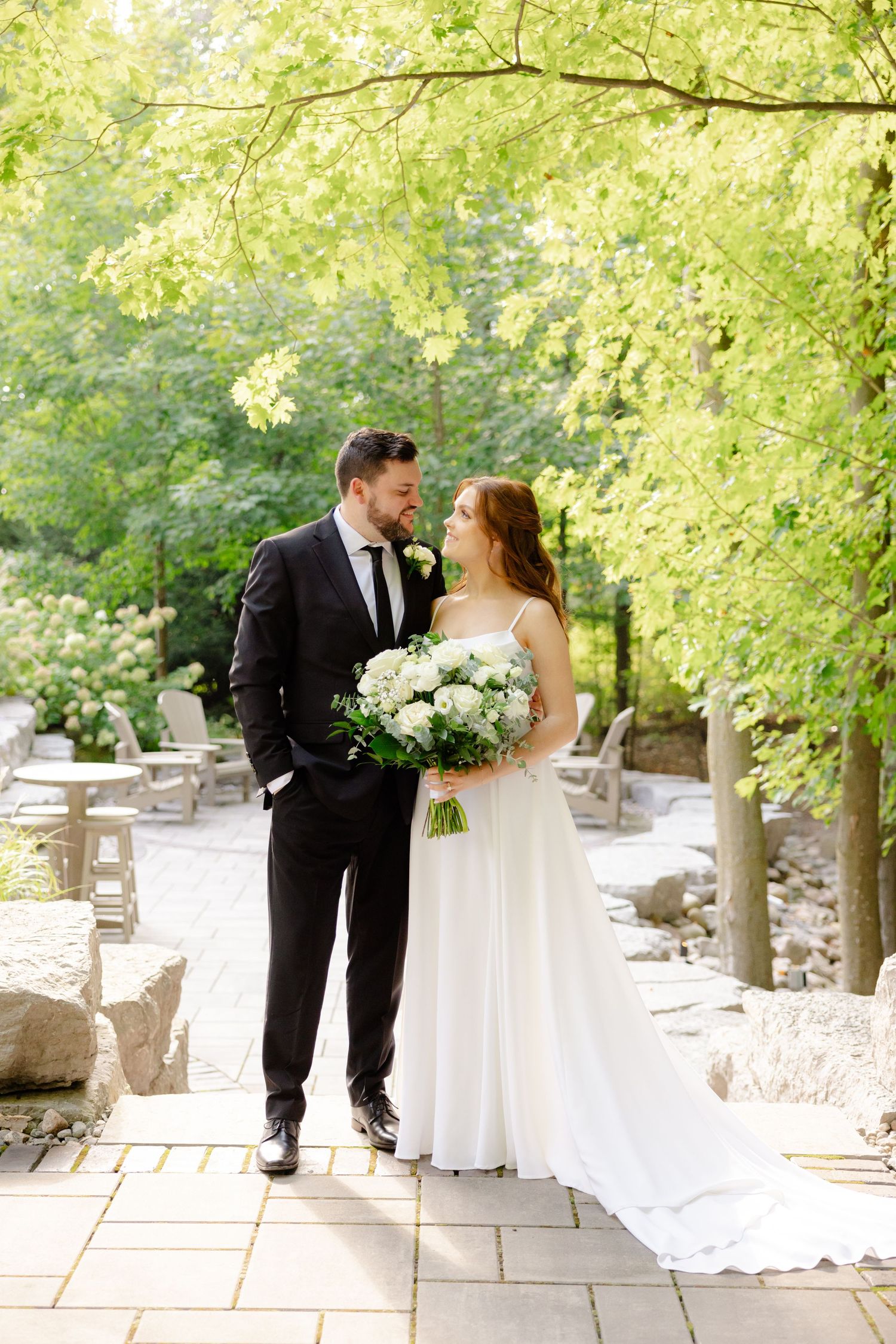 A joyful wedding couple stands on a stone path under lush green trees with a beautiful white floral bouquet.