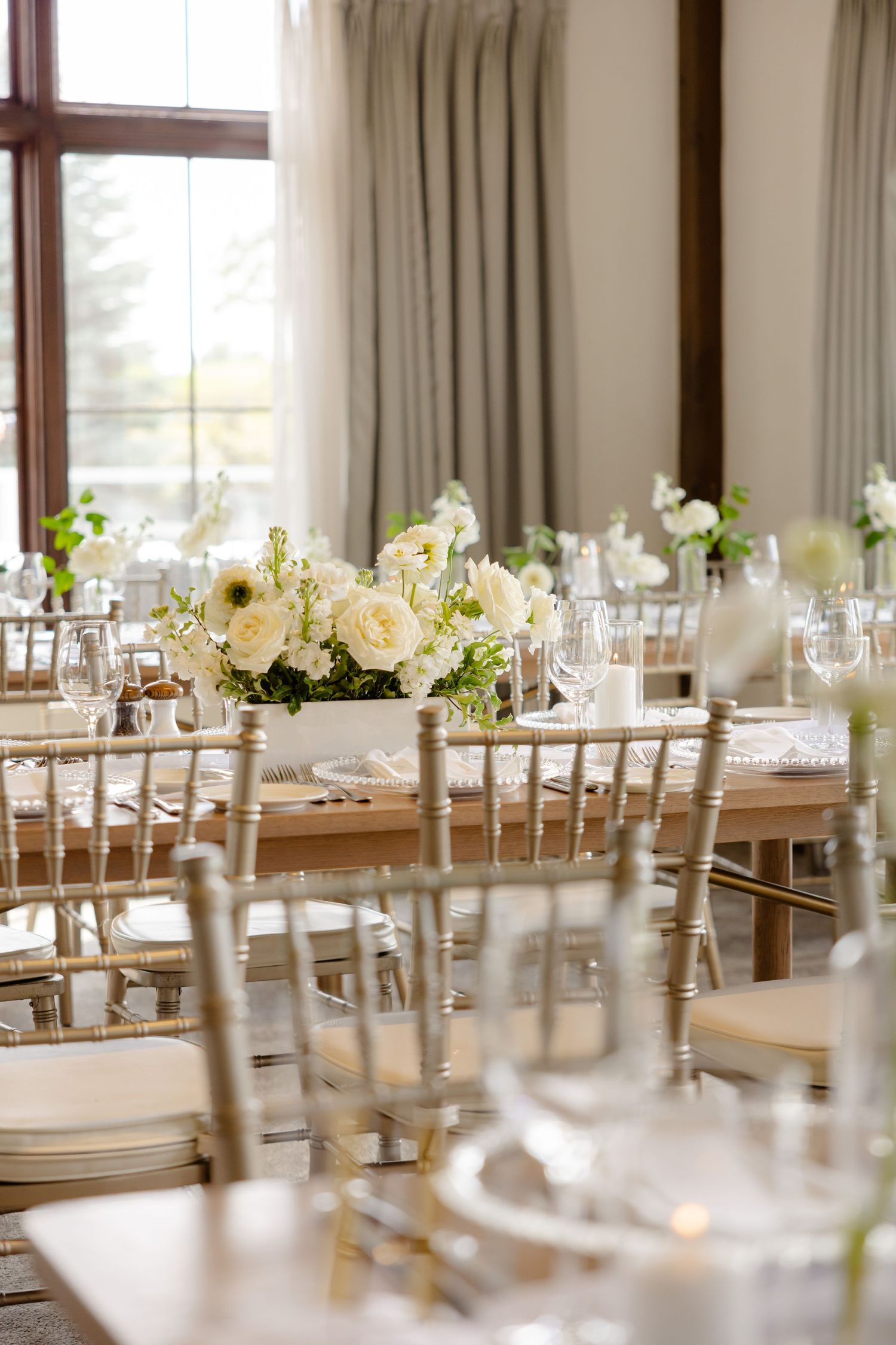 Elegant wedding reception table setup with white flower centerpieces and gold chiavari chairs in a bright venue.