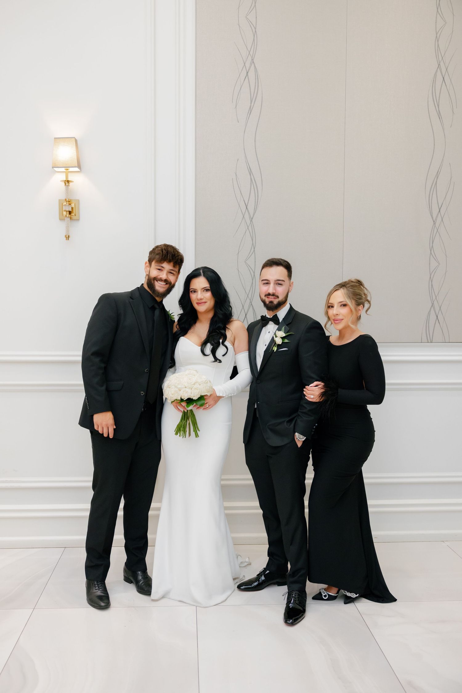 Elegant wedding party in black and white formal wear posing against light wall with decorative sconce.