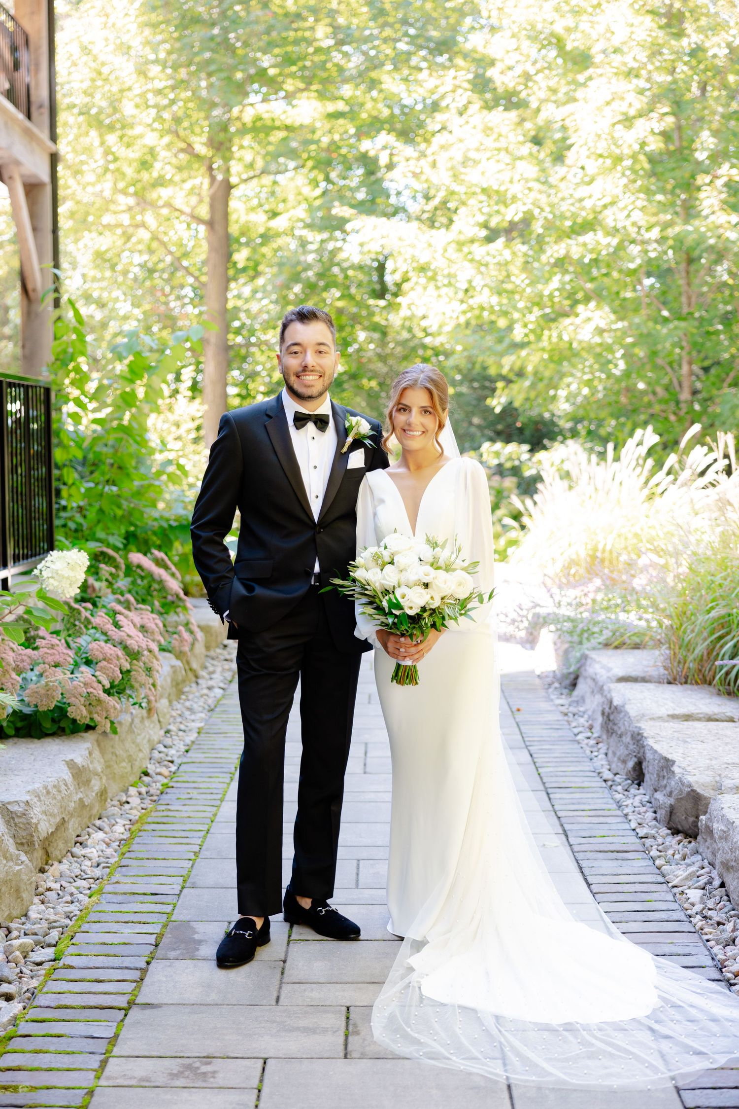 Elegant wedding portrait on stone path with natural garden backdrop and rustic architecture.