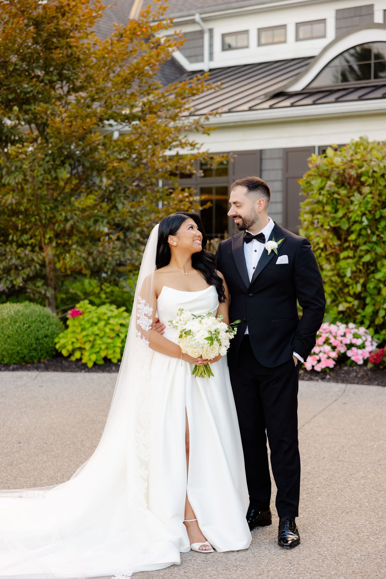 A wedding couple strolls together on a paved walkway with autumn foliage behind them.